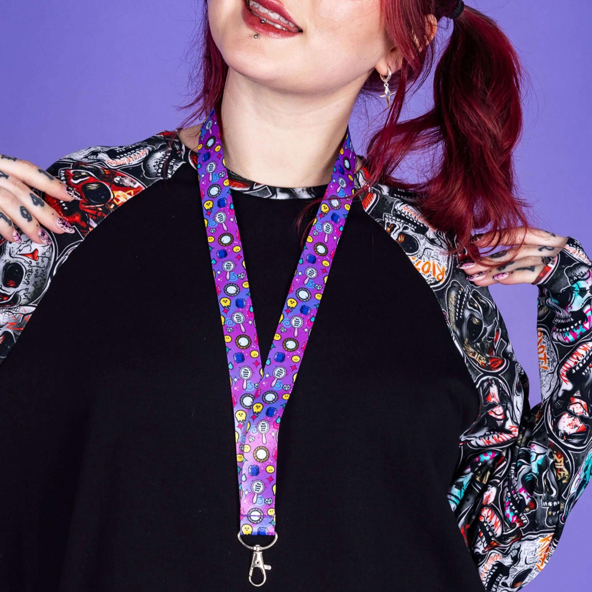 You Don't Look Sick Lanyard worn by a tattooed alternative model with red hair in bunches wearing an emo skull raglan top from wildemode. Silver clip purple lanyard features melting yellow smiley faces, mirrors, gemstones, teardrops, sparkles, swirls and dots with a centre hand held mirror reading 'you don't look sick'. The hand drawn design is raising awareness for invisible illnesses.