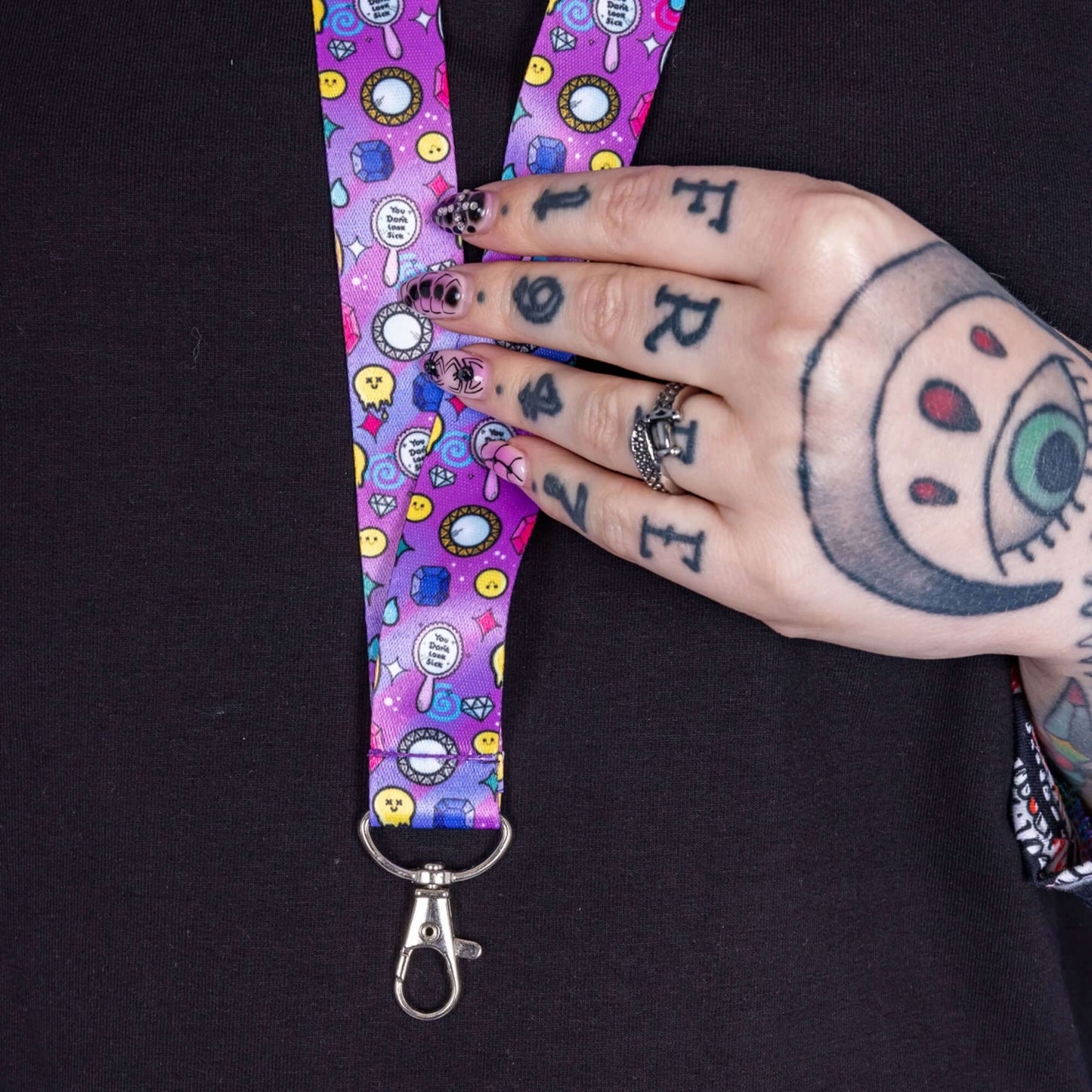 You Don't Look Sick Lanyard worn by a tattooed model with red hair. Silver clip purple lanyard features melting yellow smiley faces, mirrors, gemstones, teardrops, sparkles, swirls and dots with a centre hand held mirror reading 'you don't look sick'. The hand drawn design is raising awareness for invisible illnesses.