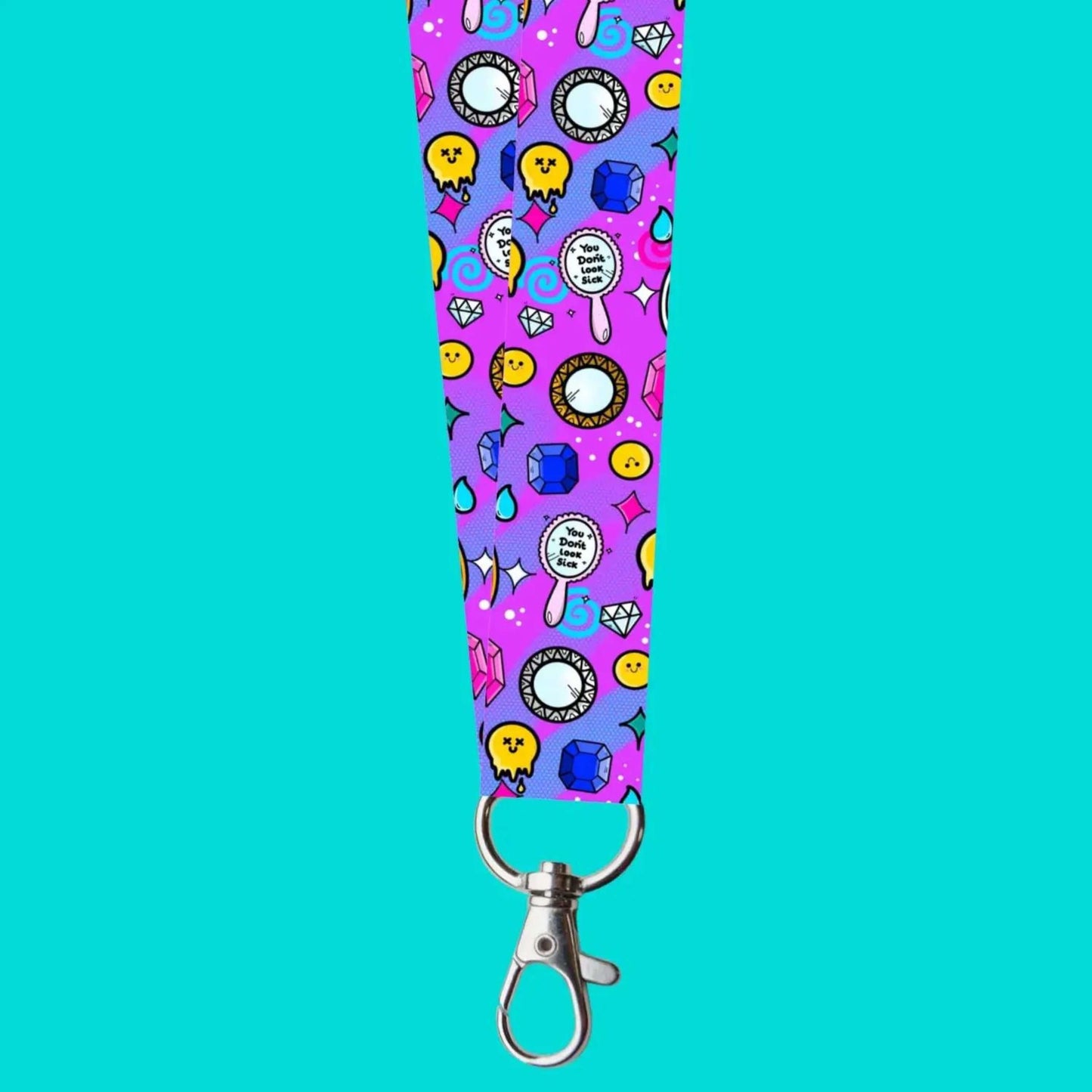 The You Don't Look Sick Lanyard on a blue background. Silver clip purple lanyard features melting yellow smiley faces, mirrors, gemstones, teardrops, sparkles, swirls and dots with a centre hand held mirror reading 'you don't look sick'. The hand drawn design is raising awareness for invisible illnesses.