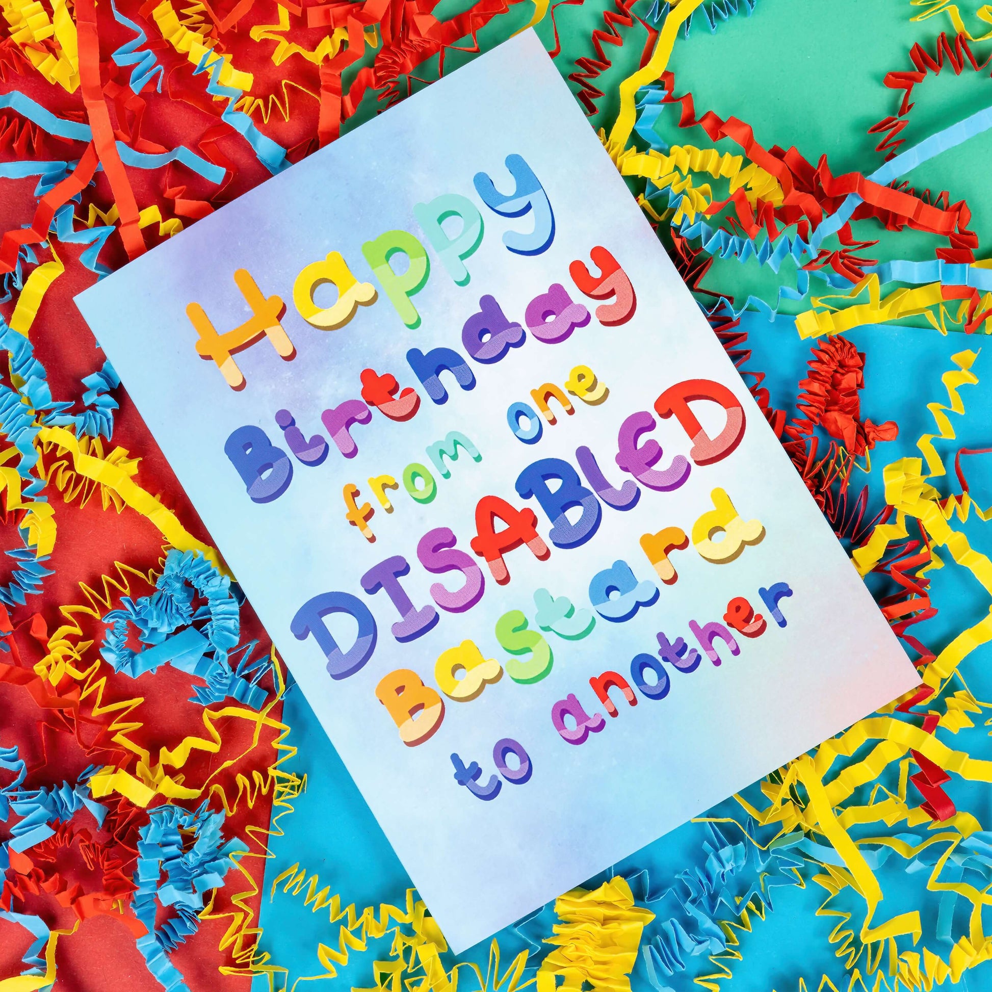 The Happy Birthday From One Disabled Bastard To Another Card on a red, blue and green background with red, yellow and blue crinkle card confetti. The pastel blue and purple a6 birthday card has rainbow bubble writing reading 'happy birthday from one disabled bastard to another'. The hand drawn design is a humorous greeting card raising awareness for disabled people.