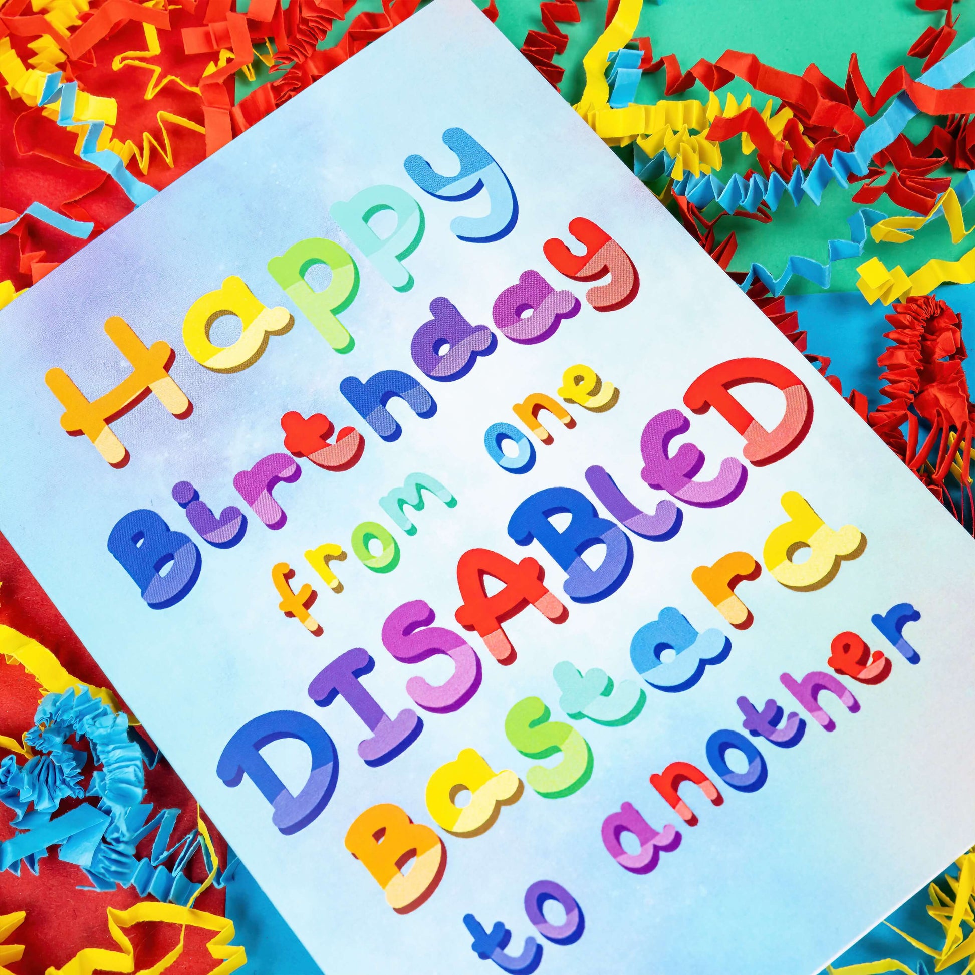 The Happy Birthday From One Disabled Bastard To Another Card on a red, blue and green background with red, yellow and blue crinkle card confetti. The pastel blue and purple a6 birthday card has rainbow bubble writing reading 'happy birthday from one disabled bastard to another'. The hand drawn design is a humorous greeting card raising awareness for disabled people.