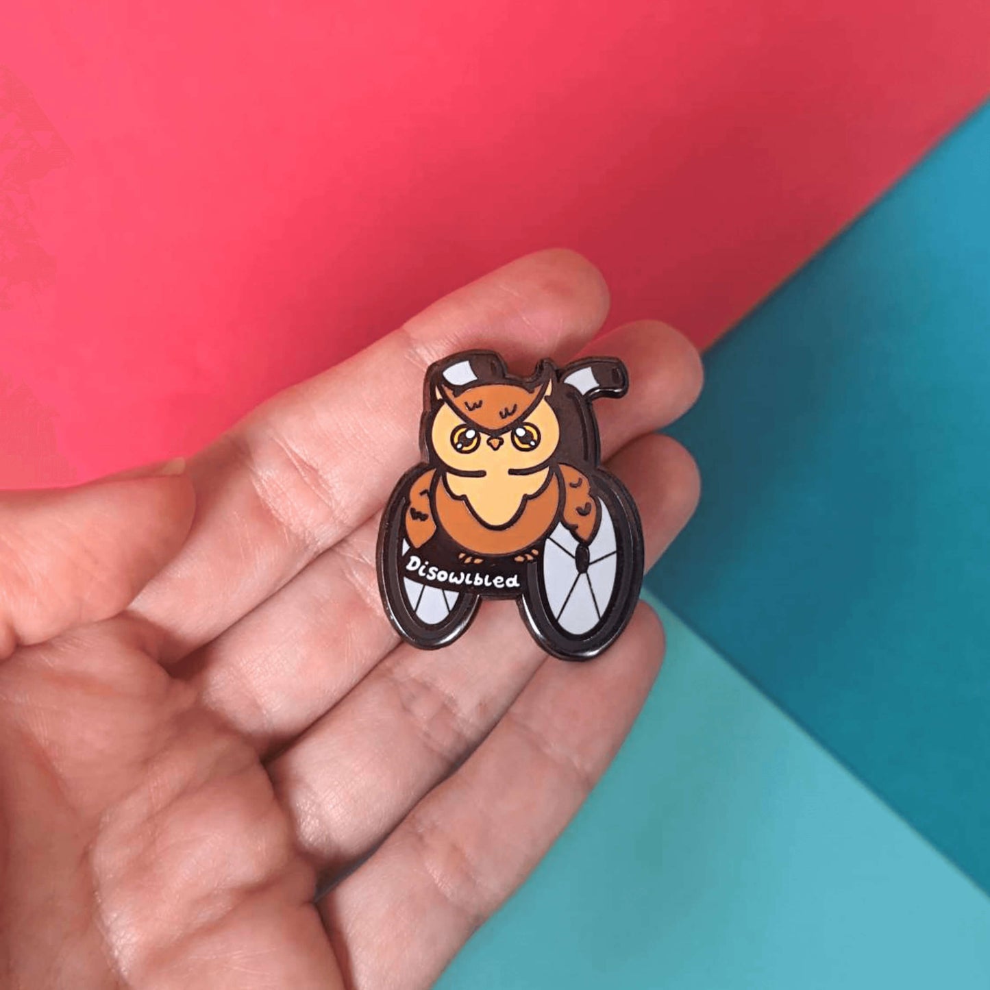 The Disowlbled Enamel Pin - Disabled being held over a red and blue background. A brown tawny owl with yellow eyes sat on a silver and black wheelchair that has white text reading 'disowlbled'. The design is raising awareness for disabilities and invisible illnesses.