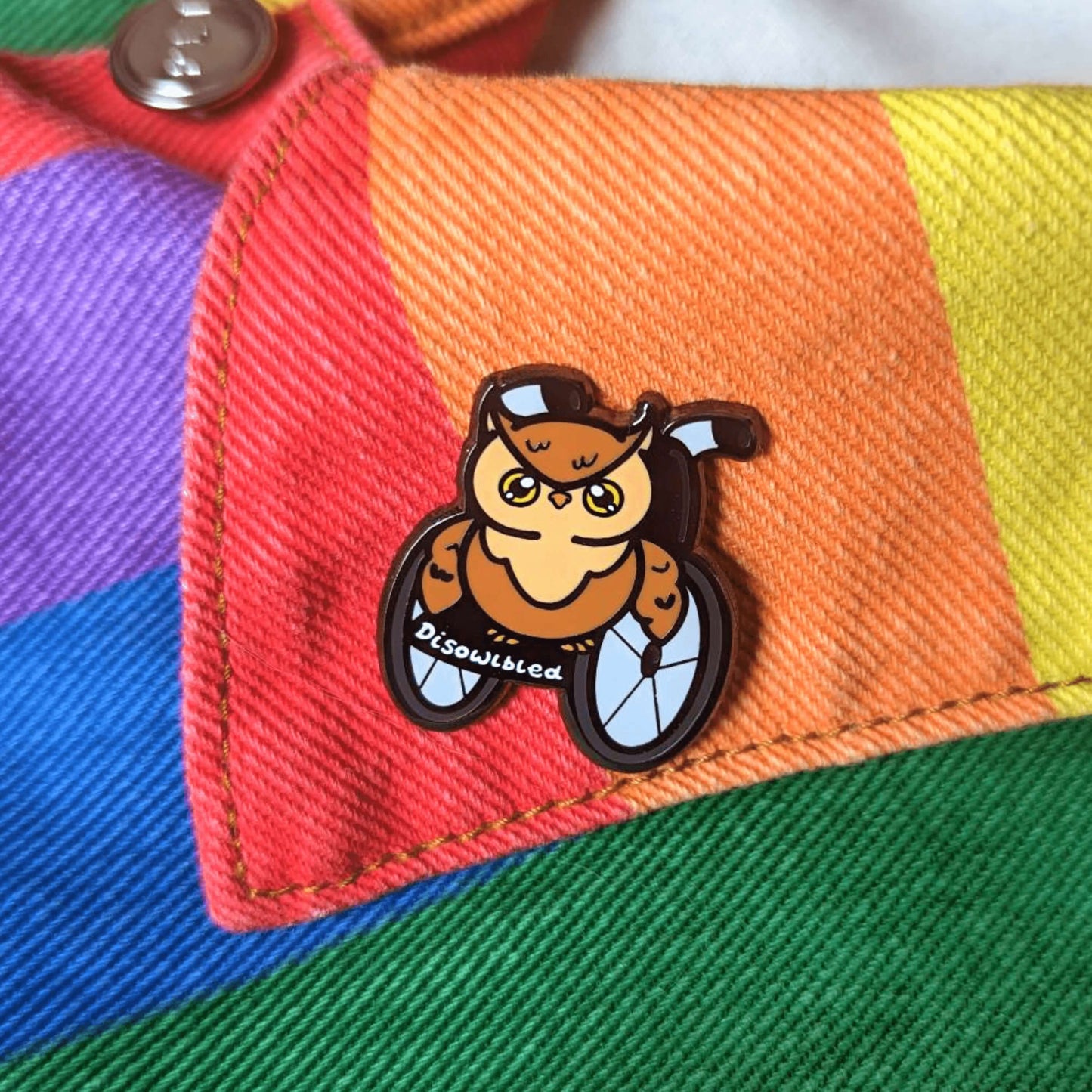 The Disowlbled Enamel Pin - Disabled on the collar of a rainbow denim jacket. A brown tawny owl with yellow eyes sat on a silver and black wheelchair that has white text reading 'disowlbled'. The design is raising awareness for disabilities and invisible illnesses.