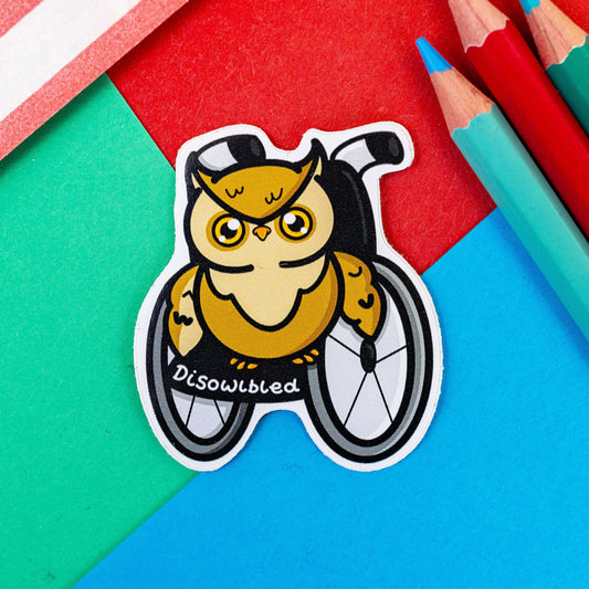 The Disowlbled Sticker - Disabled on a red, blue and green background with colouring pencils and a red stripe candy bag. The sticker of a brown tawny owl with yellow eyes sat on a silver and black wheelchair has white text reading 'disowlbled'. The design is raising awareness for disabilities and invisible illnesses.