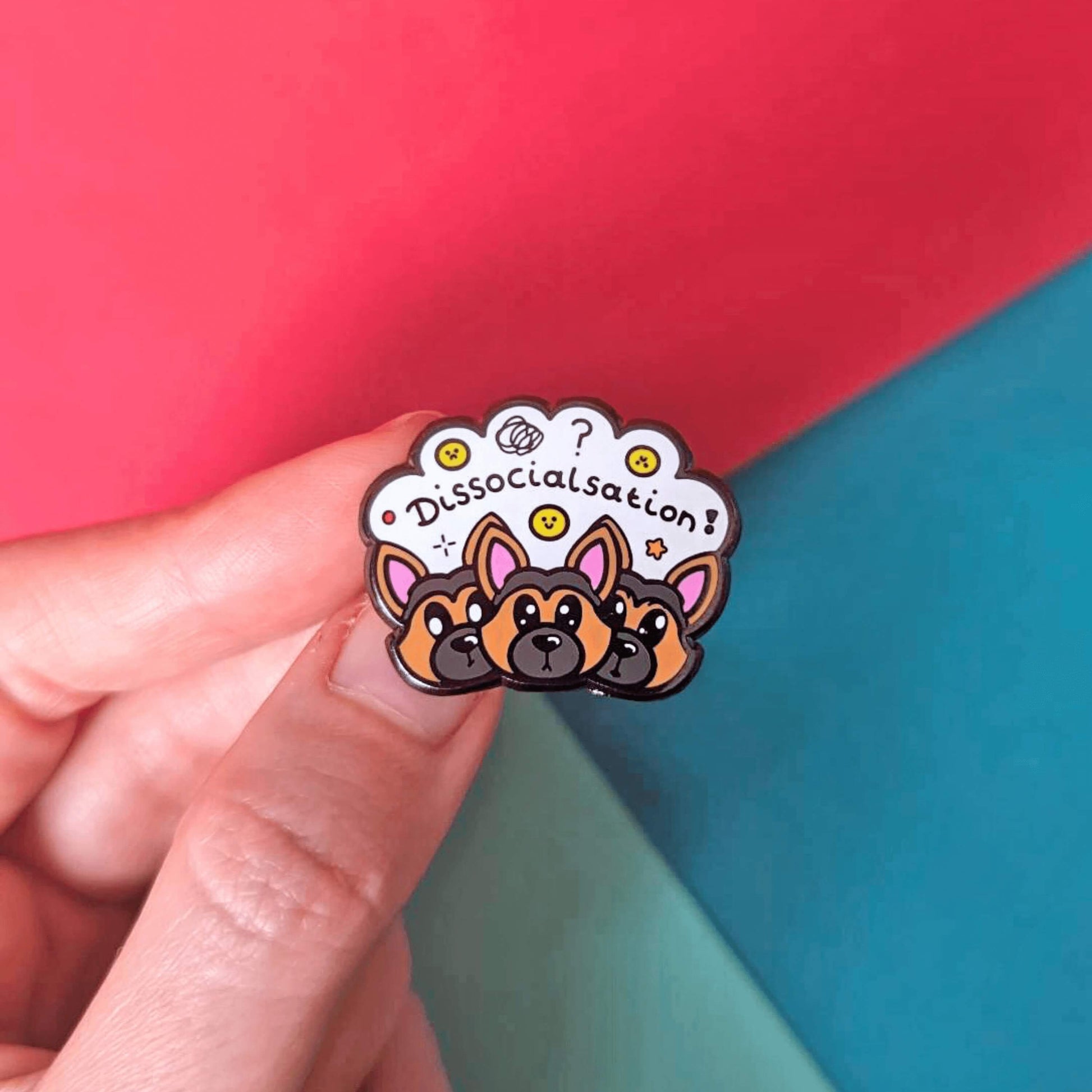 The Dissocialsation Enamel Pin - Dissociation held over a red and blue background. Three confused brown and black Alsatian dog heads with their ears perked up, above them is a white cloud with sad and happy yellow faces, sparkles, question marks and black text reading 'dissocialsation!'. The design is raising awareness for dissociation.