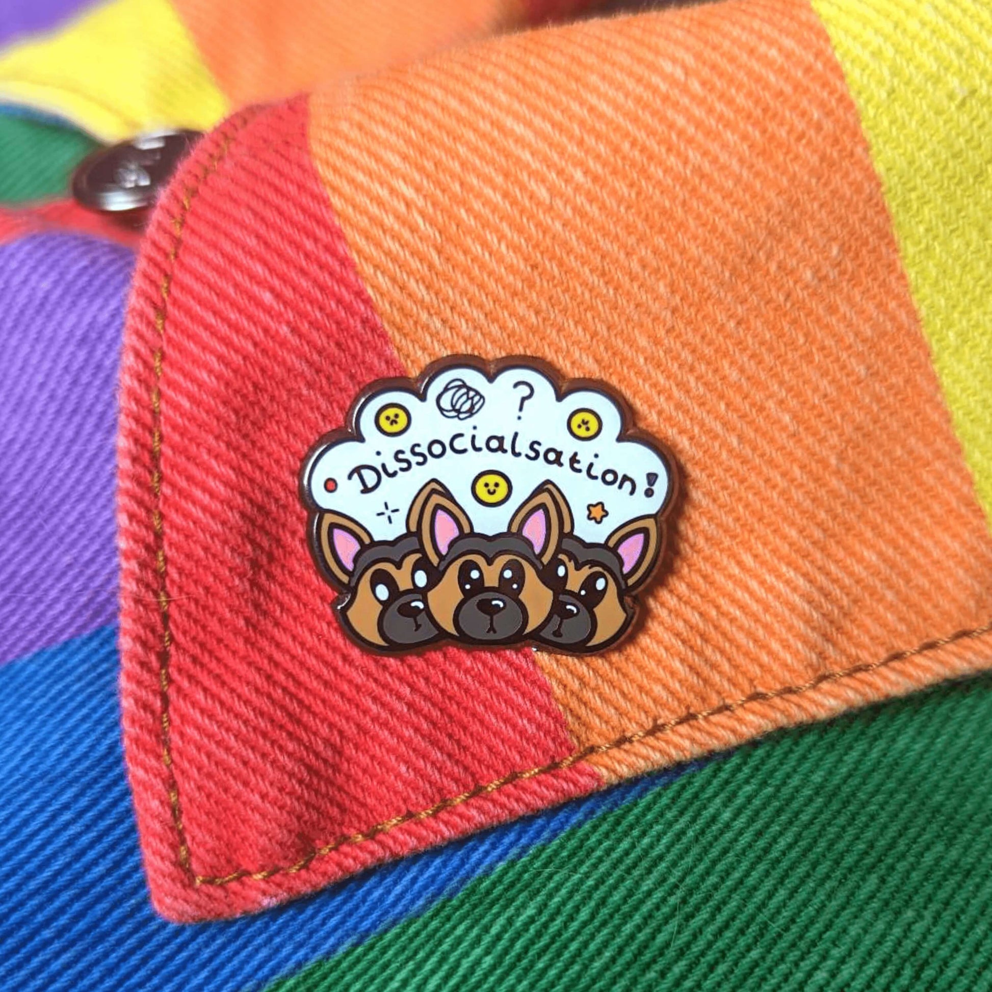 The Dissocialsation Enamel Pin - Dissociation on a rainbow denim jacket collar. Three confused brown and black Alsatian dog heads with their ears perked up, above them is a white cloud with sad and happy yellow faces, sparkles, question marks and black text reading 'dissocialsation!'. The design is raising awareness for dissociation.