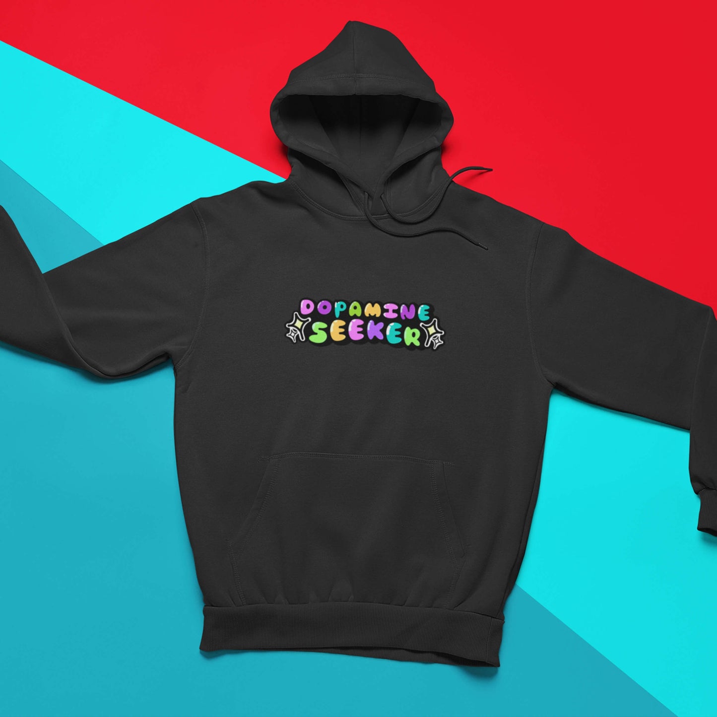 The Dopamine Seeker Black Hoodie laying on a blue and red background. The black cotton hoodie features centre rainbow bubble text that reads 'dopamine seeker' with yellow and black sparkles surrounding it. The hoodie has black drawstrings and a large centre pocket. The hoodie was designed to raise awareness for ADHD and neurodivergence. 