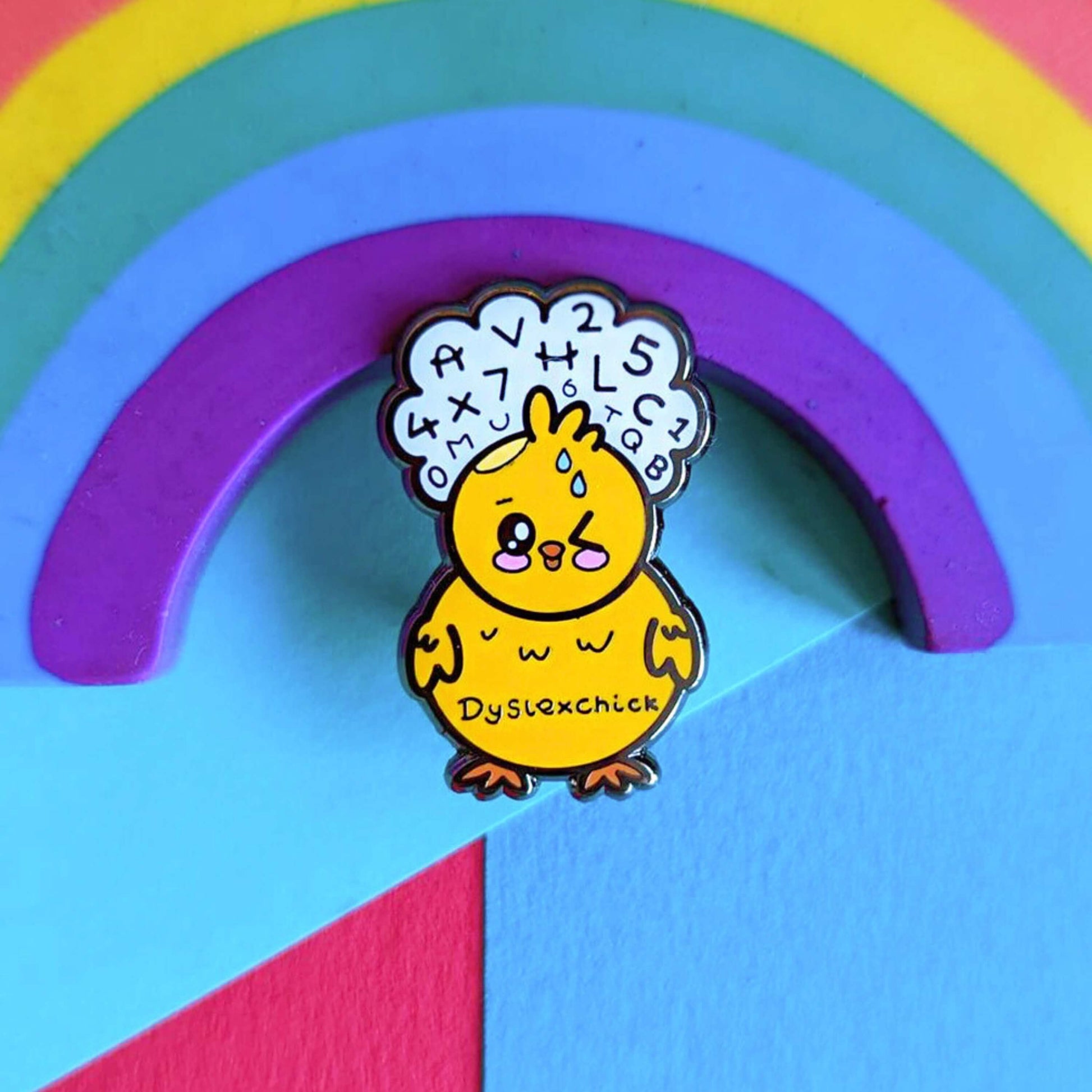 The Dyslexchick Enamel Pin - Dyslexia leaning on a rainbow on a blue background. A yellow confused chick shaped pin badge with a thought bubble above its head full of letters and numbers with 'dyslexchick' written across its middle. The hand drawn design is raising awareness for dyslexia.