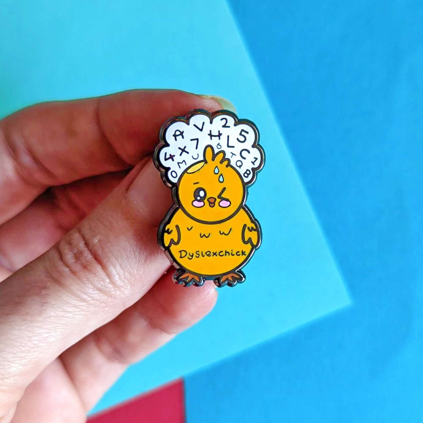 The Dyslexchick Enamel Pin - Dyslexia being held over a red and blue background. A yellow confused chick shaped pin badge with a thought bubble above its head full of letters and numbers with 'dyslexchick' written across its middle. The hand drawn design is raising awareness for dyslexia.