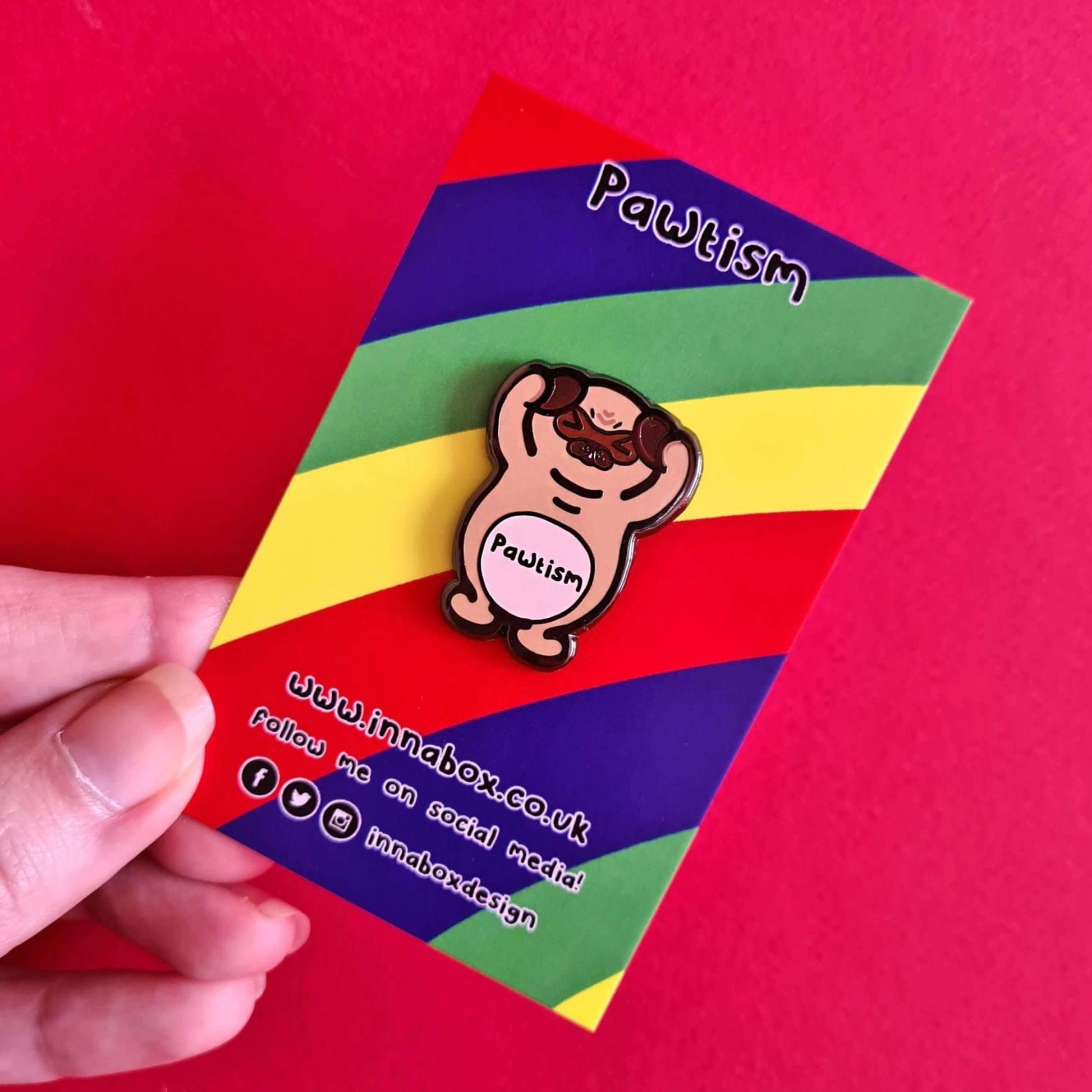 Pawtism Enamel Pin - Autism on rainbow backing card held by a hand on a red background. The enamel pin is of a brown pug with its hands on its ears and the word pawtism written across its belly. The enamel pin is to raise awareness for neurodiversity and autism.