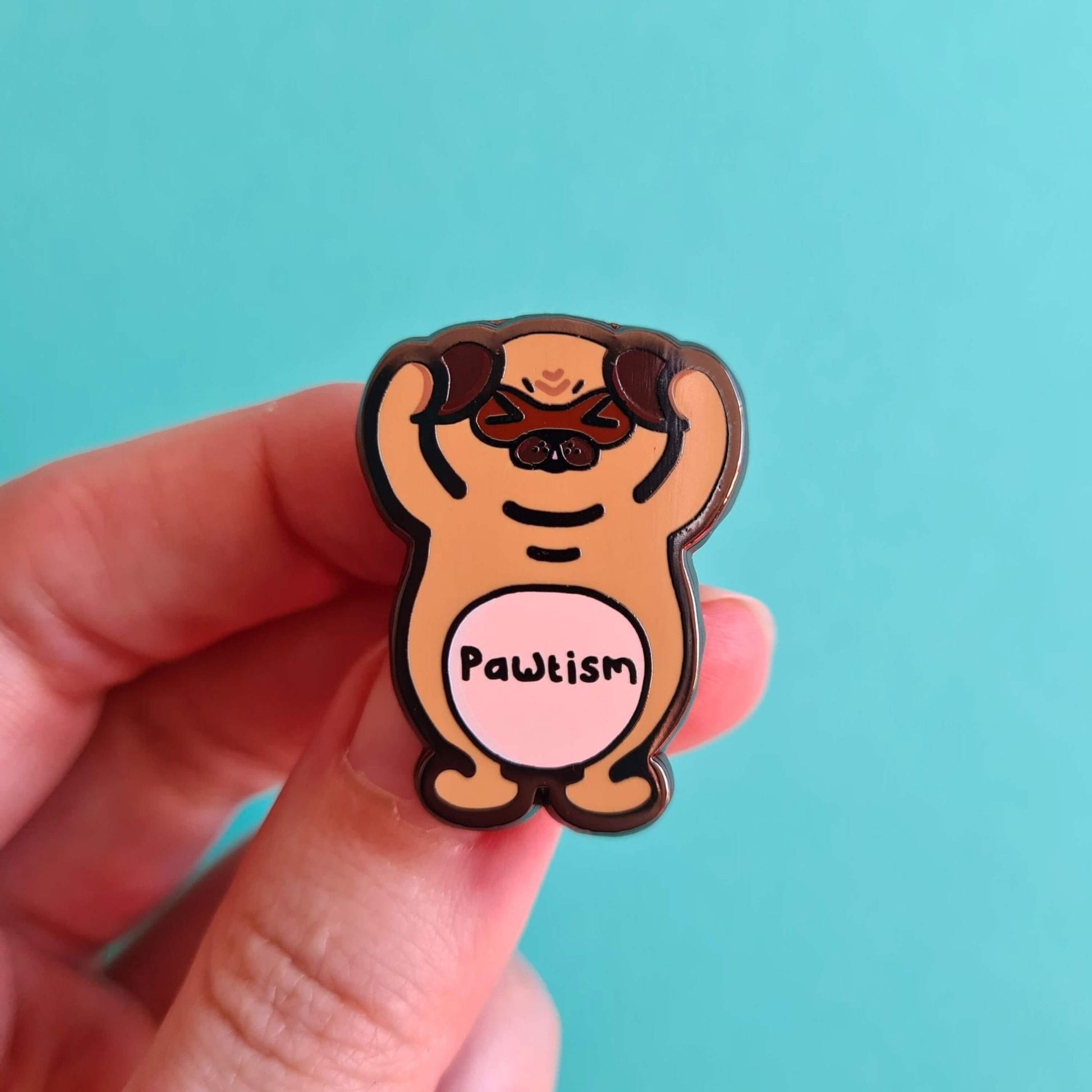Pawtism Enamel Pin - Autism on a blue background with a hand holding it. The enamel pin is of a brown pug with its hands on its ears and the word pawtism written across its belly. The enamel pin is to raise awareness for neurodiversity and autism.