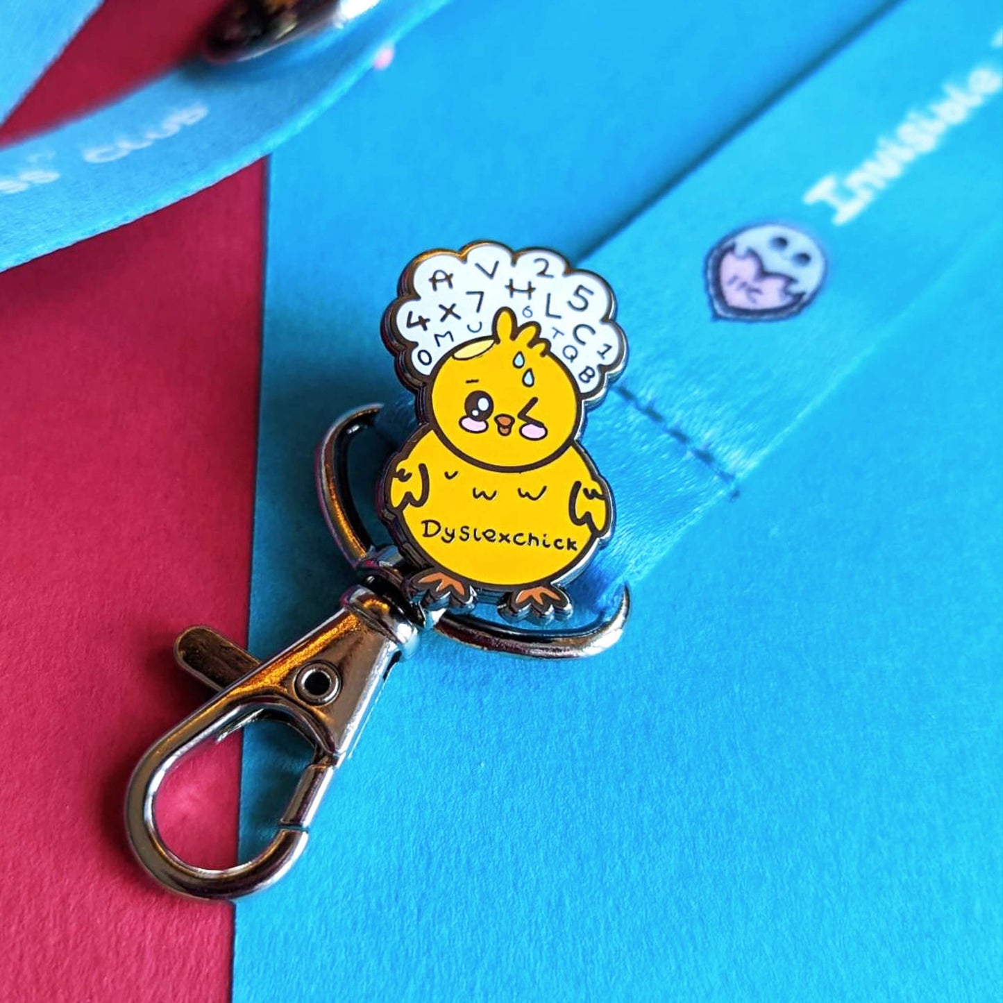 The Dyslexchick Enamel Pin - Dyslexia pinned to an innabox invisible illness blue lanyard with silver lobster clip on a red and blue background. A yellow confused chick shaped pin badge with a thought bubble above its head full of letters and numbers with 'dyslexchick' written across its middle. The hand drawn design is raising awareness for dyslexia.