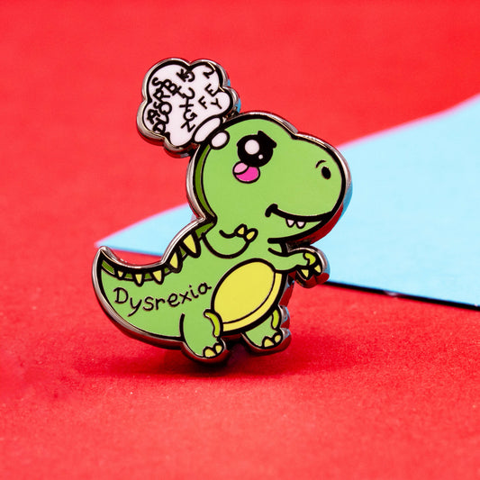 A close-up of the Innabox Dysrexia Dinosaur enamel pin showing the green dinosaur with pink cheeks, a yellow belly, and a thought bubble filled with jumbled letters. The word “Dysrexia” is written on its side. Shot on a red surface with a pop of blue background, celebrating hidden disabilities.