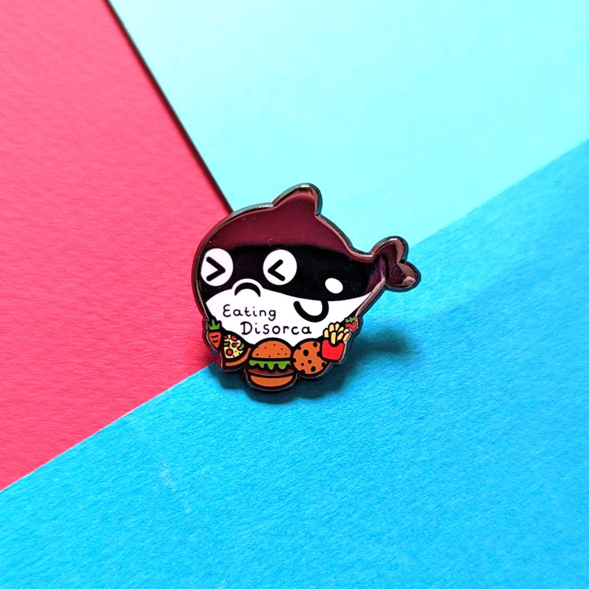 The Eating Disorca Orca Whale Enamel Pin - Eating Disorder on a red and blue background. The black orca whale shaped enamel pin has a stressed expression whilst surrounded by burgers, pizzas, cookies, fries and fruit with 'eating disorca' across its middle. The design is raising awareness for eating disorders.