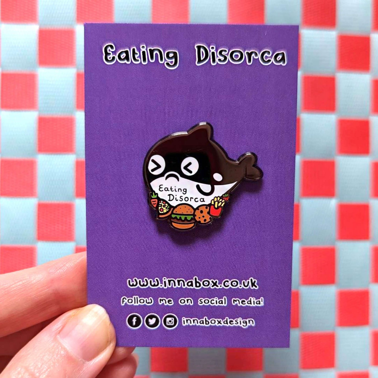 The Eating Disorca Orca Whale Enamel Pin - Eating Disorder on purple backing card with 'eating disorca' written above in black and innabox website and social media handles below, held over a red and white background. The black orca whale shaped enamel pin has a stressed expression whilst surrounded by burgers, pizzas, cookies, fries and fruit with 'eating discorca' across its middle. The design is raising awareness for eating disorders.