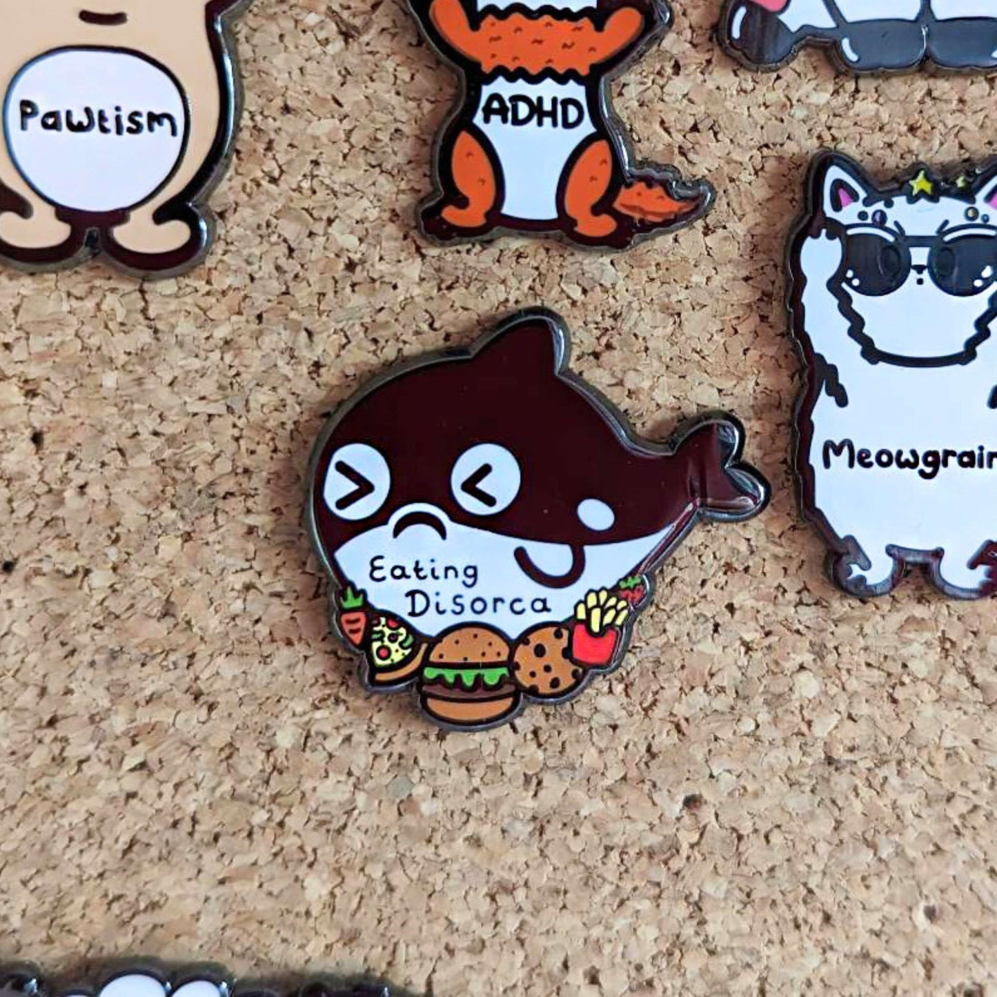 The Eating Disorca Orca Whale Enamel Pin - Eating Disorder pinned on a corkboard with other pin badges from innabox. The black orca whale shaped enamel pin has a stressed expression whilst surrounded by burgers, pizzas, cookies, fries and fruit with 'eating discorca' across its middle. The design is raising awareness for eating disorders.