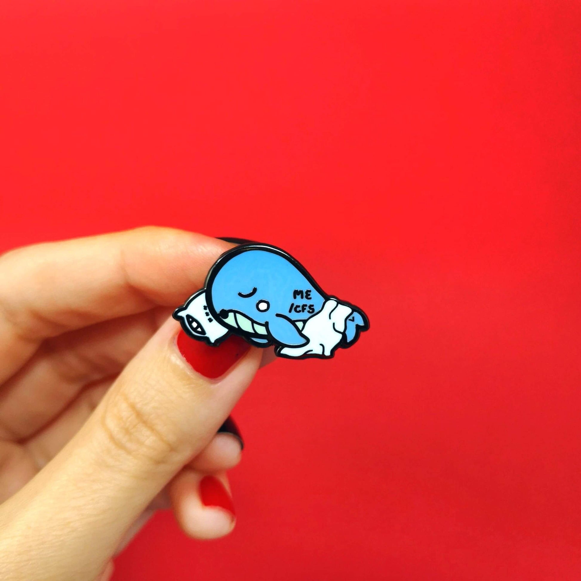Mywhalegic Enamel Pin - Myalgic Encephalomyelitis (ME/CFS) held by a hand on a red background. The enamel pin is a sleeping blue whale laying on a white pillow with a white blanket over it and ME/CFS written on its back. The enamel pin is designed to raise awareness for Myalgic encephalomyelitis or chronic fatigue syndrome