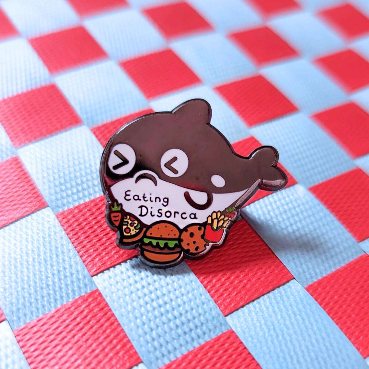 The Eating Disorca Orca Whale Enamel Pin - Eating Disorder on a red and white background. The black orca whale shaped enamel pin has a stressed expression whilst surrounded by burgers, pizzas, cookies, fries and fruit with 'eating discorca' across its middle. The design is raising awareness for eating disorders.