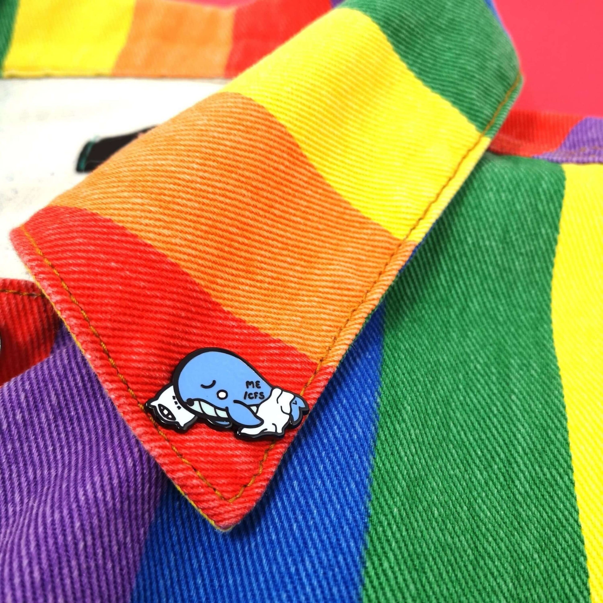 Mywhalegic Enamel Pin - Myalgic Encephalomyelitis (ME/CFS) pinned onto a rainbow jacket. The enamel pin is a sleeping blue whale laying on a white pillow with a white blanket over it and ME/CFS written on its back. The enamel pin is designed to raise awareness for Myalgic encephalomyelitis or chronic fatigue syndrome