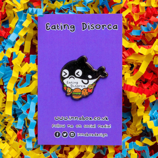 The Eating Disorca Orca Whale Enamel Pin - Eating Disorder on purple backing card with 'eating disorca' written above in black and innabox website and social media handles below laid on red, yellow and blue card confetti. The black orca whale shaped enamel pin has a stressed expression whilst surrounded by burgers, pizzas, cookies, fries and fruit with 'eating discorca' across its middle. The design is raising awareness for eating disorders.
