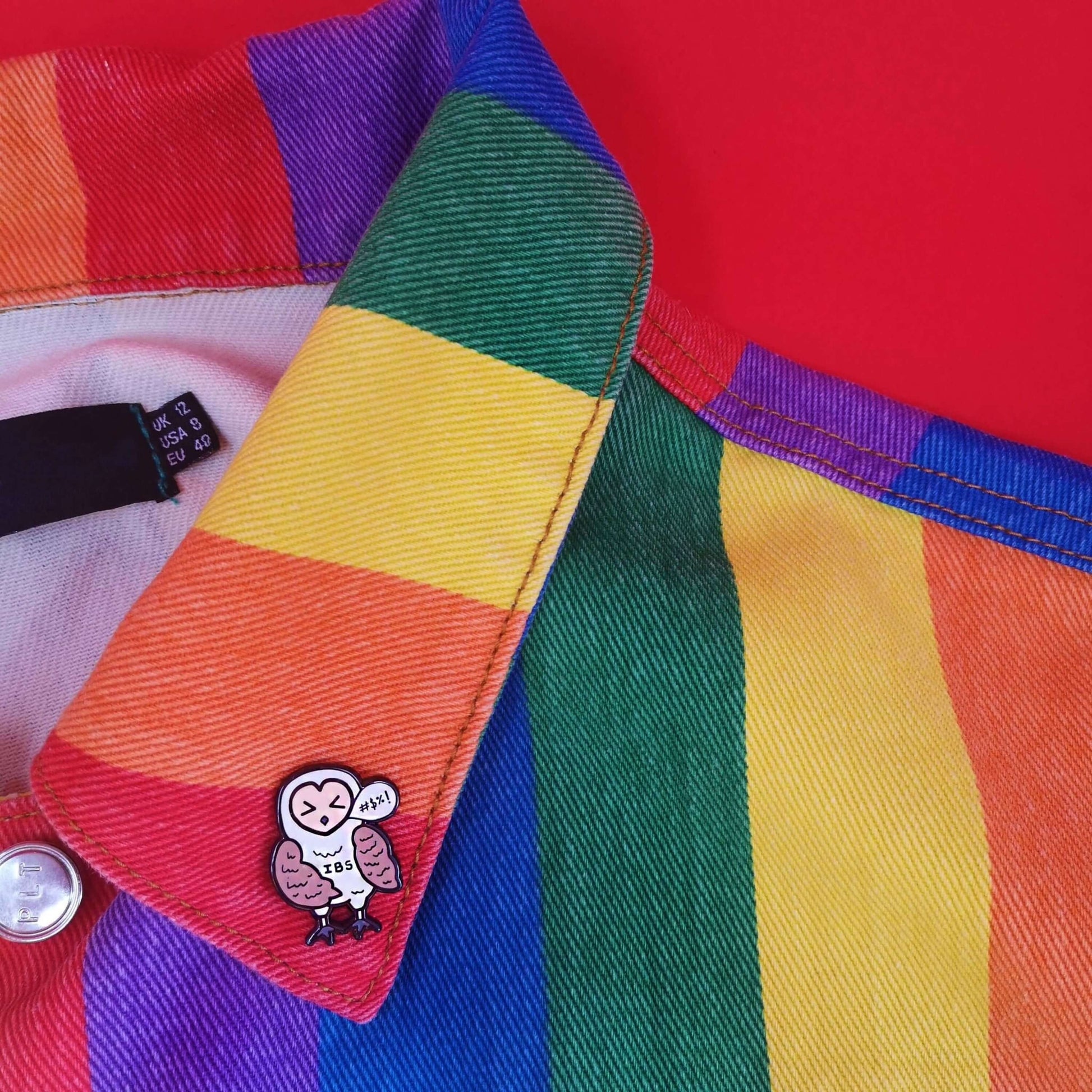 Irritable Owl Syndrome Enamel Pin - Irritable bowel syndrome (IBS) pinned to a rainbow jacket. The enamel pin is of a barn owl with a sweary text speech bubble and has its eyes shut with the letters IBS written on its belly. Hand drawn design to raise awareness for Irritable bowel syndrome (IBS)