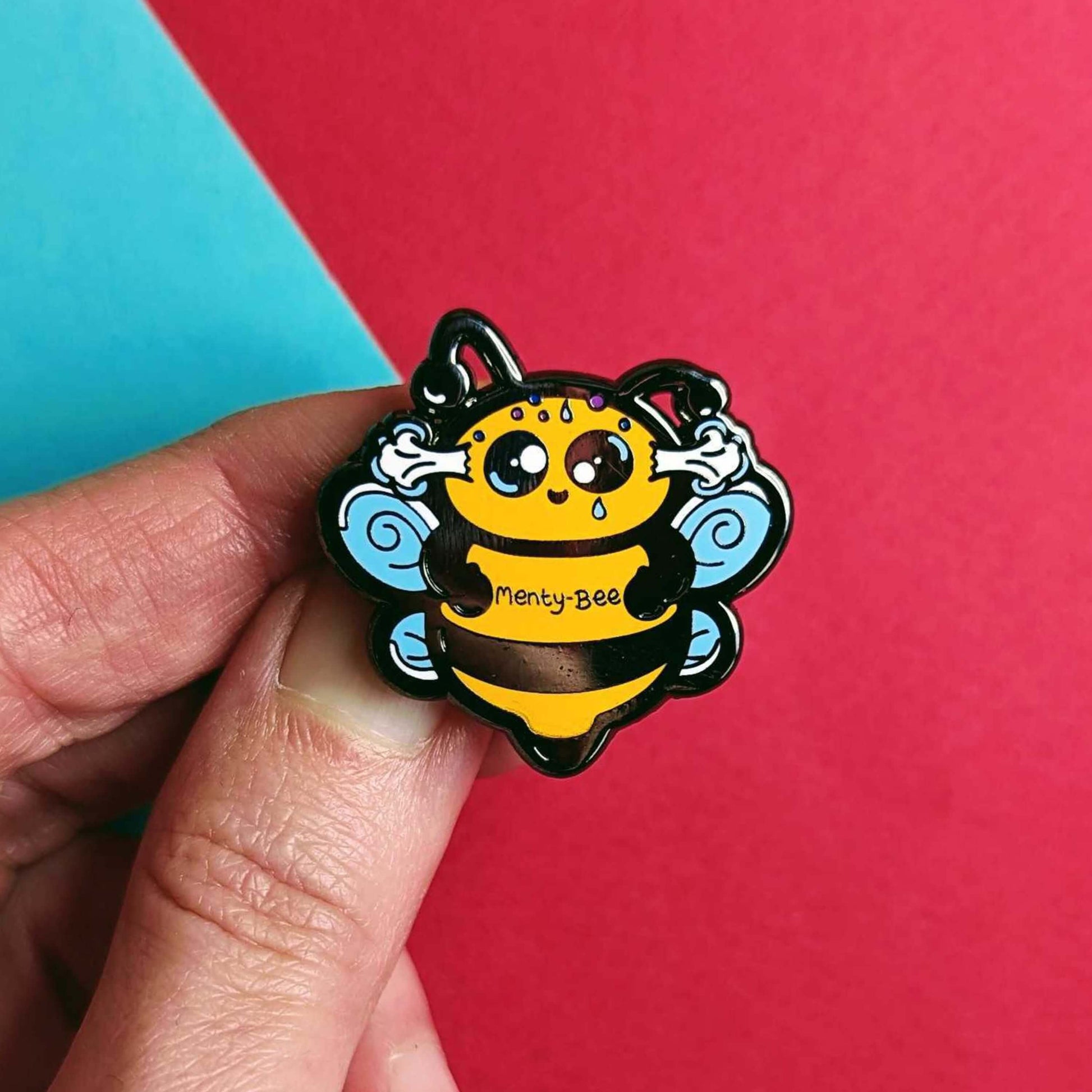 Menty-Bee Enamel Pin - Mental Breakdown held in front of a red and blue background. The enamel pin is of a bee looking stressed with steam coming out of its head and the text menty-Bee on its chest. Hand drawn design made to raise awareness for mental breakdowns