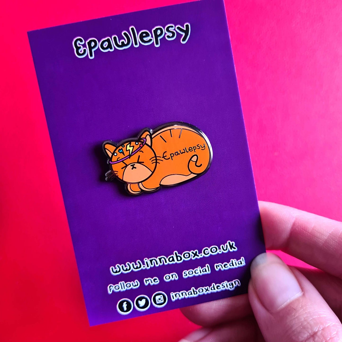Epawlepsy Enamel Pin - Epilepsy on a purple backing card red background. The enamel pin is a ginger cat with eyes scrunched closed and symbols to represent a dizzy spell drawn across his head. Epawlepsy is written on the cats stomach. Enamel pin is designed to raise awareness for epilepsy