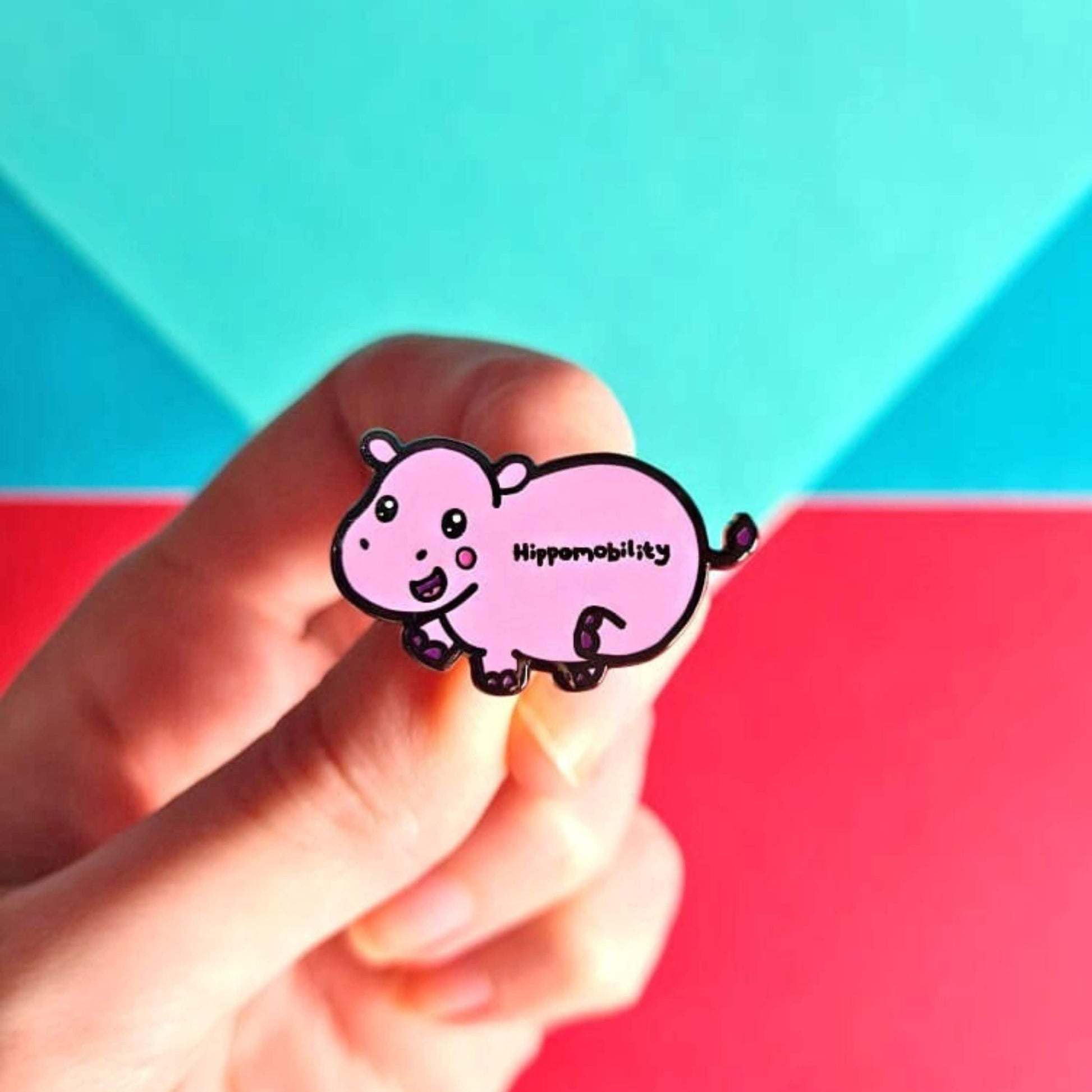 Hippmobility Enamel Pin - Hyper Mobility being held in front of a red and blue background. The enamel pin is of a pink happy hippo with the text hippomobility written on its stomach. The enamel pin is designed to raise awareness for hyper mobility