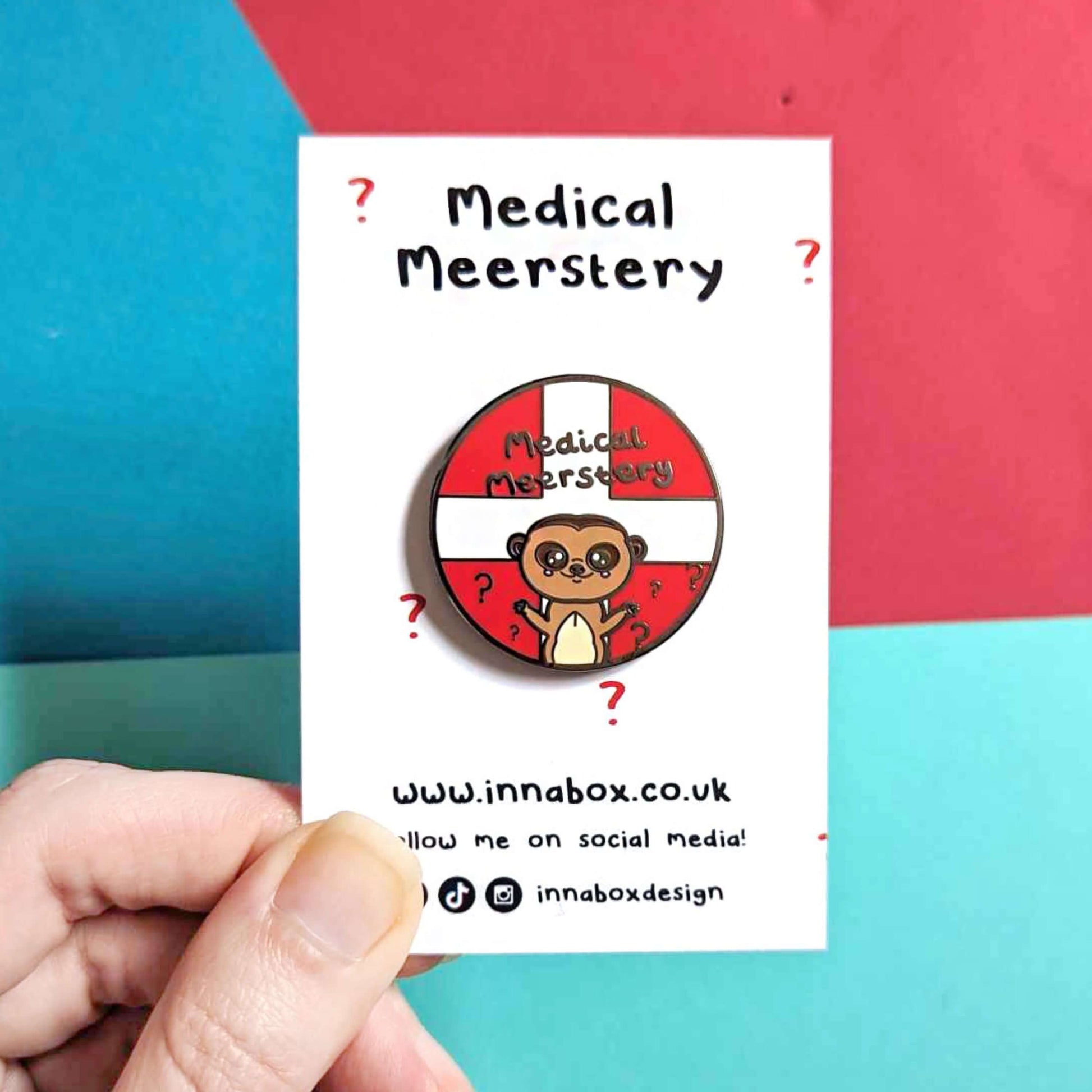 Medical Mystery Meerkat Enamel Pin on white backing card with red questions mark on held in front of a red and blue background. The enamel pin is a red circle with white first aid cross and a cute meerkat at the bottom with big eyes and has it's little arms open wide. There are black question marks around the meerkat and 'medical meerstery' written above it in black writing. Hand drawn design made to raise awareness for invisible and chronic illnesses