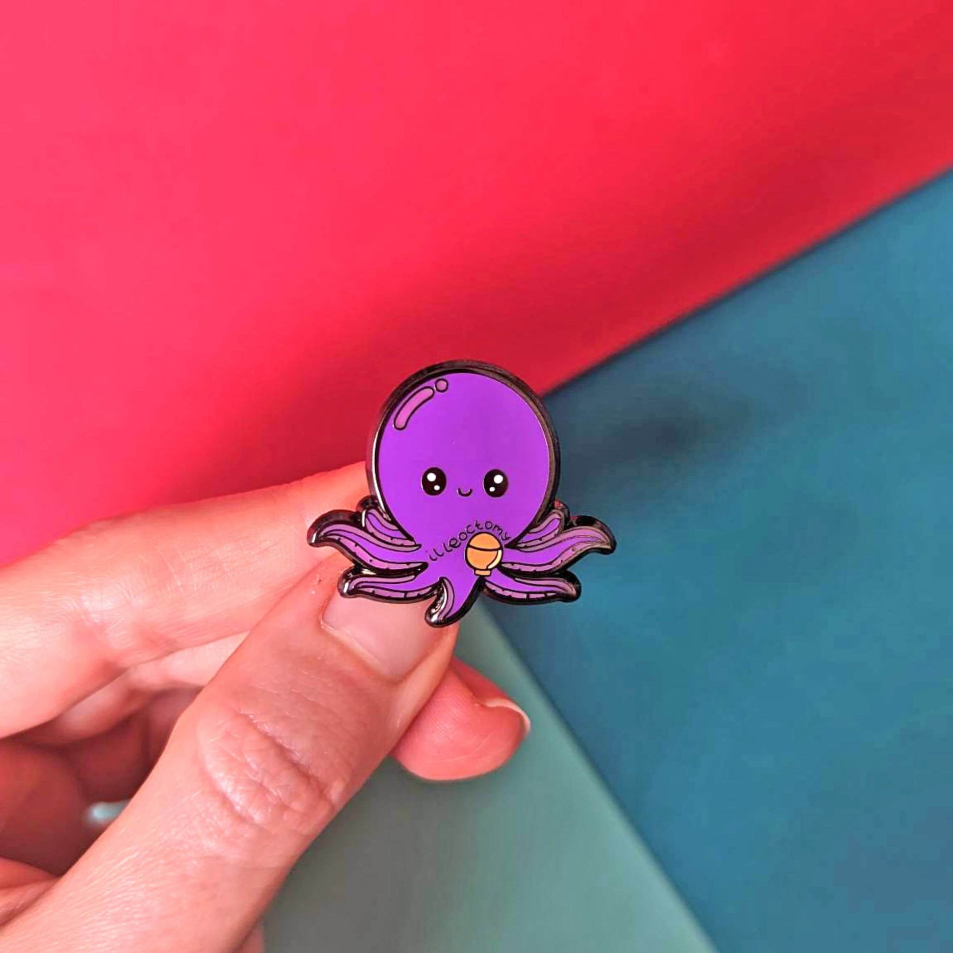 Ileoctomy Enamel Pin - Ileostomy held in front of a blue and red background. The enamel pin is a cute smiling purple octopus sticker with text saying ileoctomy on its belly with a stoma bag underneath. Enamel pin designed to raise awareness for Ileostomy