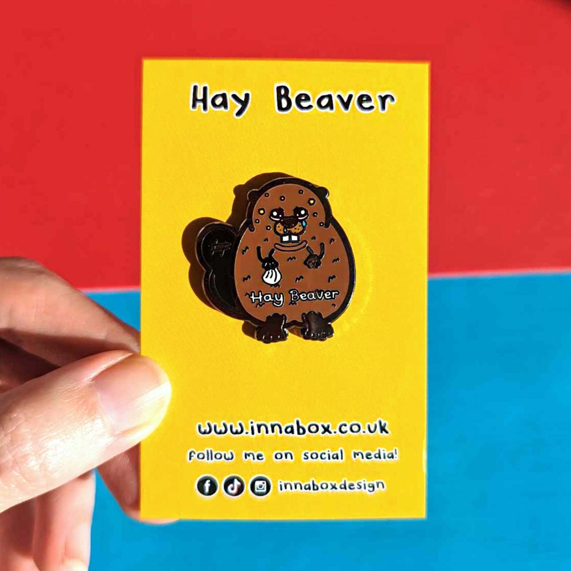 Hay Beaver Enamel Pin - Hay Fever on yellow backing card held in front of blue and red background. The enamel pin is a brown beaver with watery eyes, dripping nose and yellow spots on face and is holding a tissue with it's little hand. Hay beaver is written across its belly. The enamel pin is designed to raise awareness for hay fever or allergic rhinitis