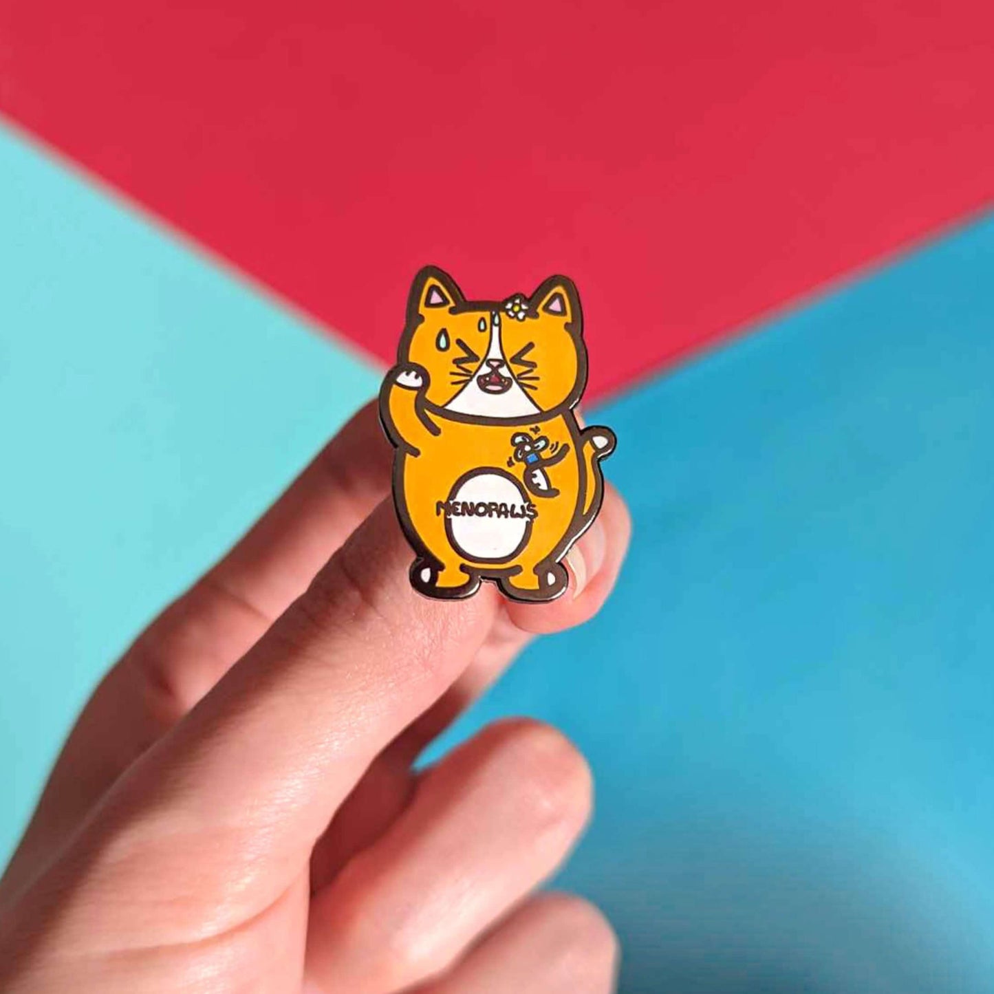 Menopaws Enamel Pin - Menopause held in front of a red and blue background. The enamel pin is of a menopausal ginger and white cat with a daisy in it's hair. The cat is holding an electric fan and is looking hot with sweat droplets on it's face. Hand drawn design made to raise awareness for menopause