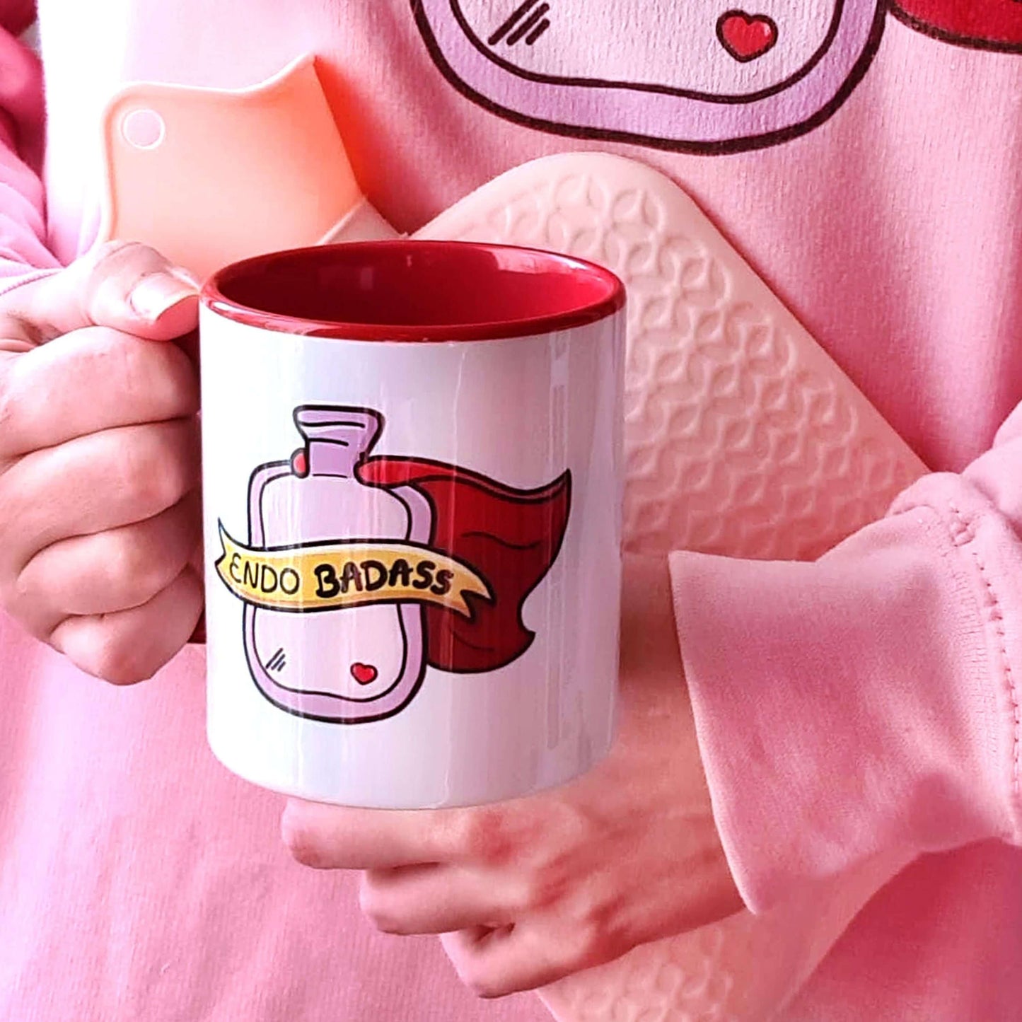 Person wearing a pink top holding a pink water bottle to their stomach and also holding the Endo Badass Mug - Endometriosis. The white mug has a red handle and inside with an illustration of a pink hot water bottle wearing a red cape. There is a yellow banner across the bottle with black text inside that reads 'endo badass'. Hand drawn design made to raise awareness for endometriosis.