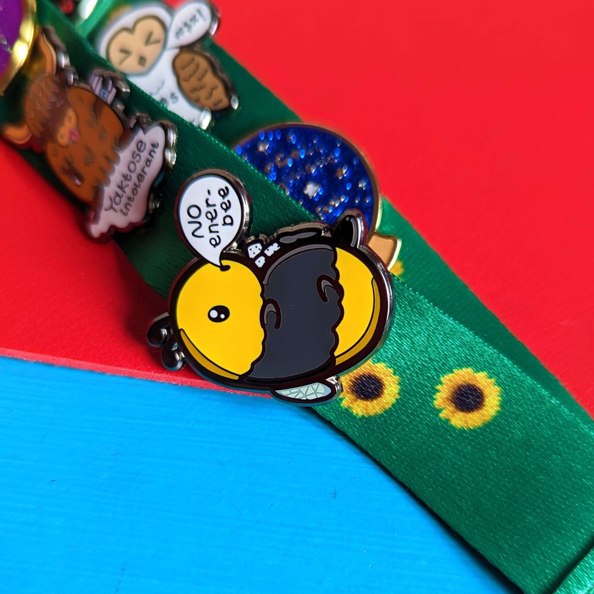 Low Energy Bee Enamel Pin pinned to a sunflower lanyard with other Innabox pins. The enamel pin is a cute yellow and black bee lying on it's wings with its arms, legs, little belly and stinger in the air. The bee has a speech bubble coming from it's mouth with 'NO ener-bee' written inside in black writing. Hand drawn design made to raise awareness for chronic fatigue.