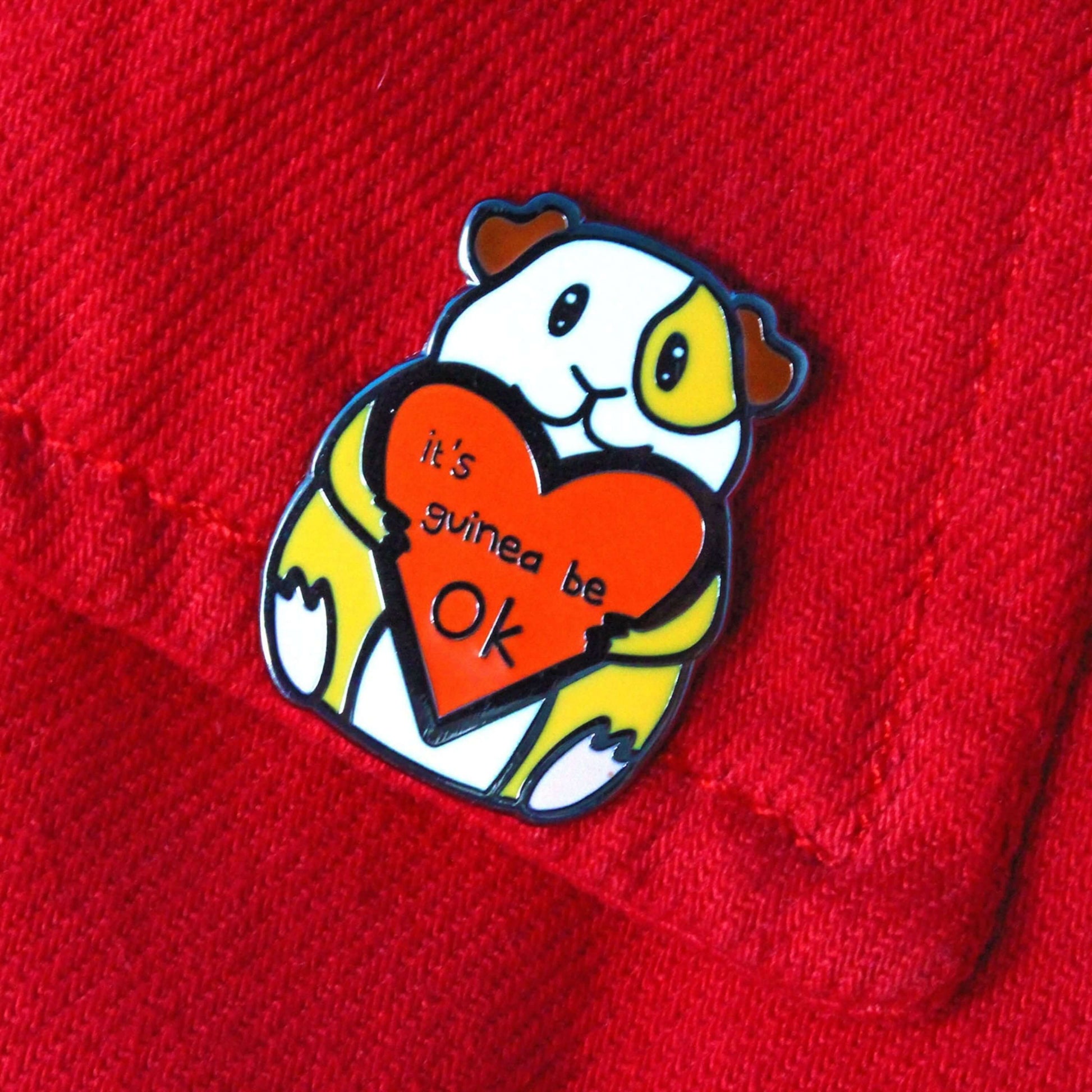 Guinea Pig Mental Health Enamel Pin pinned onto a red jacket. The enamel pin is of a smiling guinea pig holding a big red heart with the text it's guinea be ok inside. The enamel pin is deigned to raise awareness for mental health.