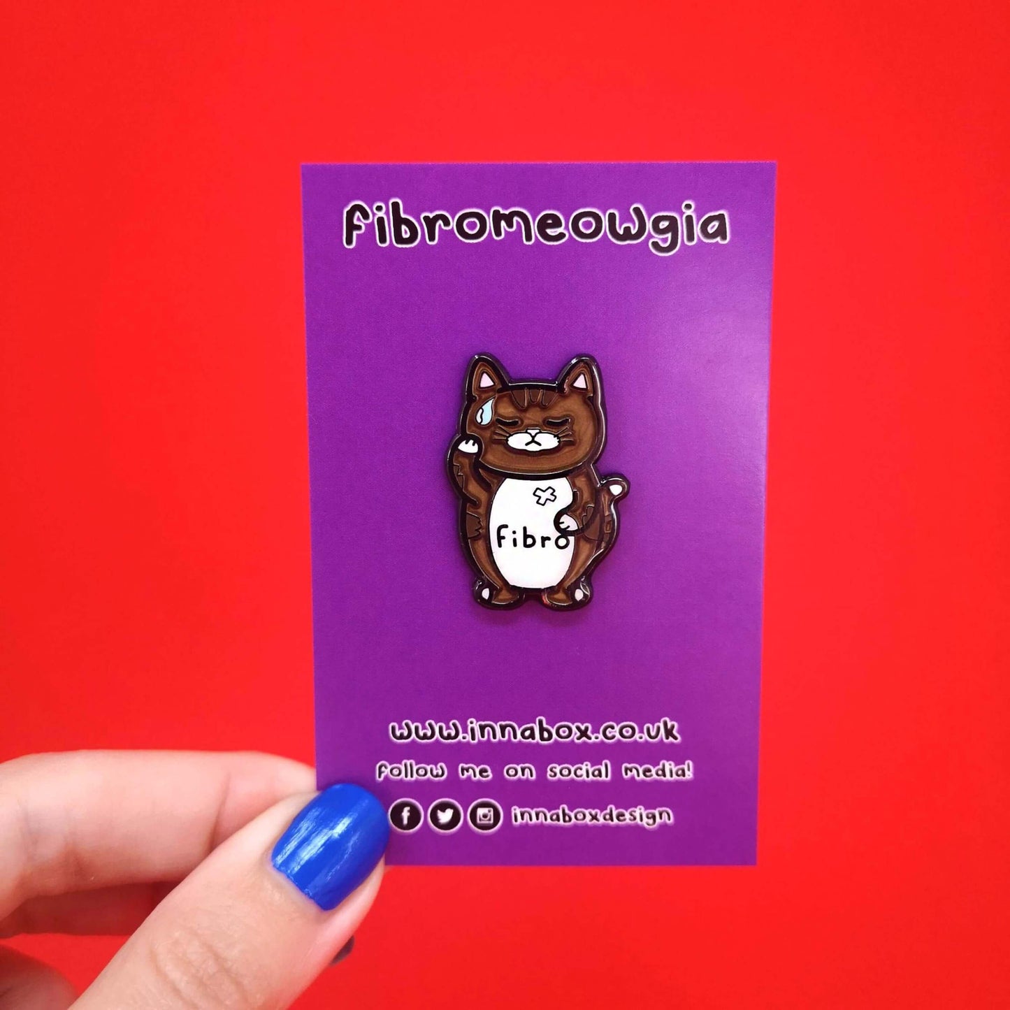 Fibromeowgia Enamel Pin - Fibromyalgia on purple backing card held in front of a red background. The enamel pin is a brown cat with a white stomach with fibro written across it. The cat has its eyes closed, a sweat droplet on its forehead and holding its head in its paw. The enamel pin is designed to raise awareness for fibromyalgia
