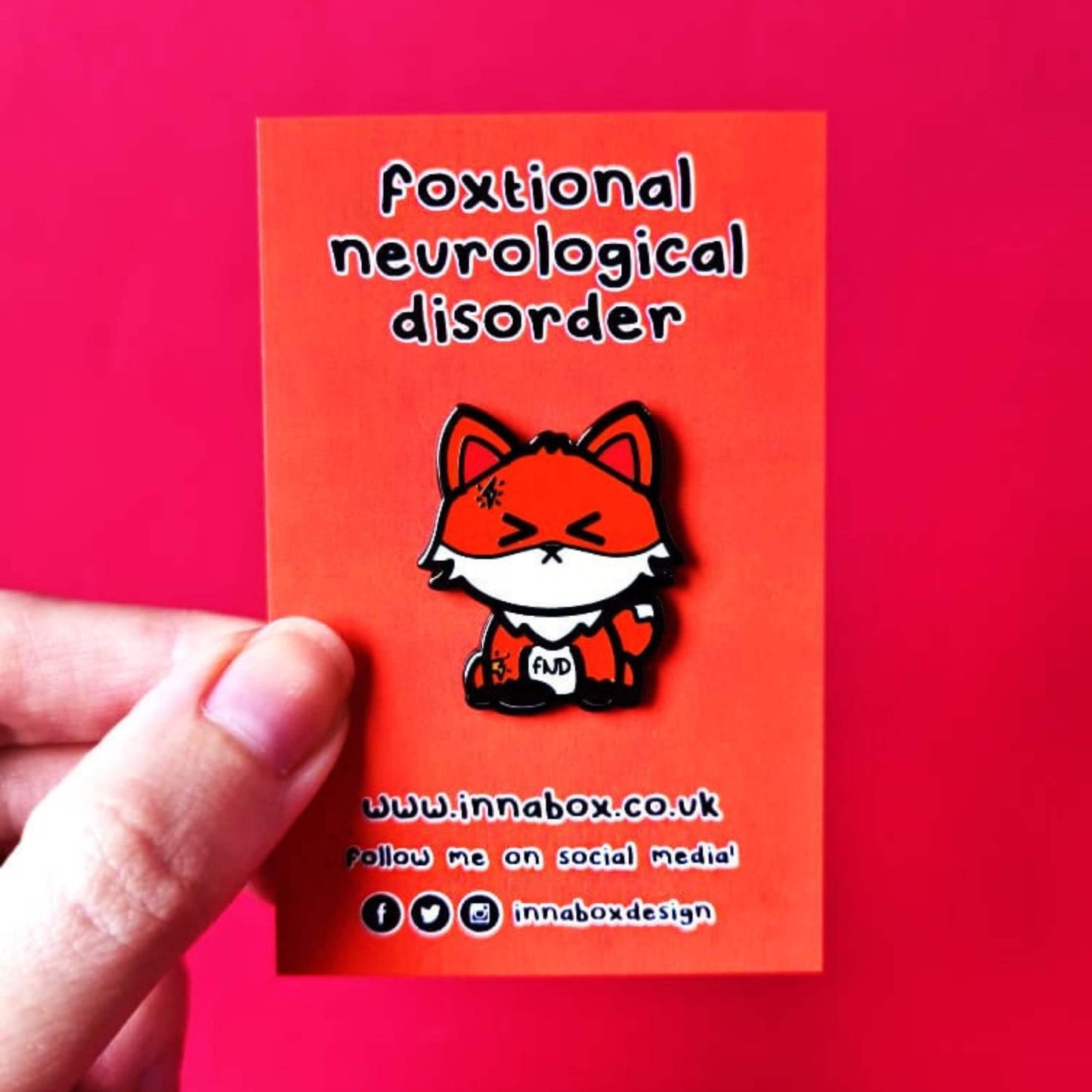 Foxtional Neurological Disorder - Functional Neurological Disorder (FND) Enamel Pin on orange backing card held in front of a red background. The enamel pin is a fox with a pained expression on its face and yellow lightning illustration on its head and arm. The enamel pin is designed to raise awareness for Functional Neurological Disorder (FND)