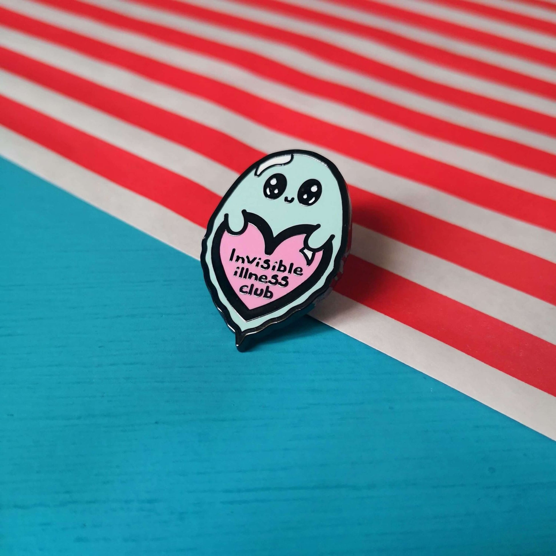 Invisible Illness Club Enamel Pin shown on a red and white striped paper bag on a blue background. The enamel pin is of a cute smiling ghost holding a pink heart with text saying invisible illness club. The enamel pin is designed to raise awareness for hidden and chronic illnesses