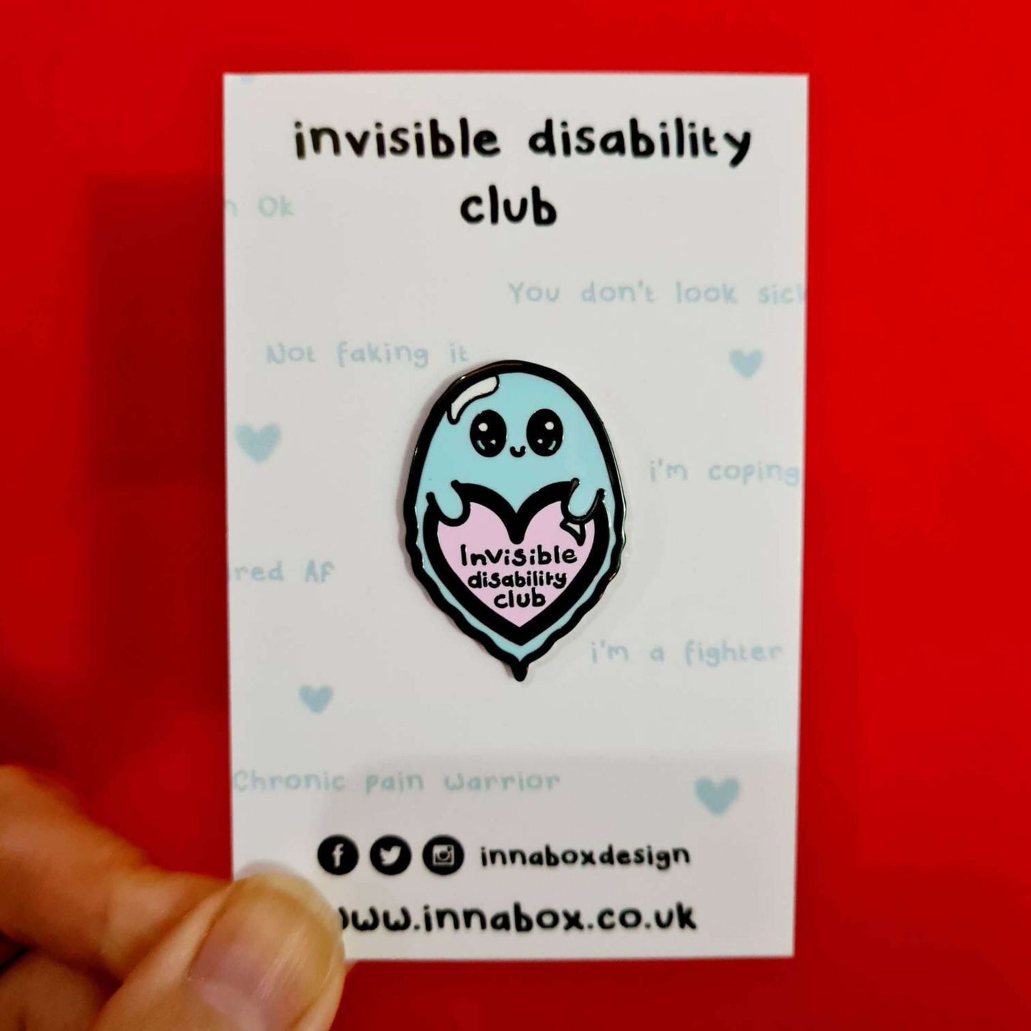 Invisible Disability Club Enamel Pin on white backing card with light blue quotes written on it in front of a red background. The enamel pin is of a cute smiling ghost holding a pink heart with text saying invisible disability club. The enamel pin is designed to raise awareness for hidden and chronic disabilities