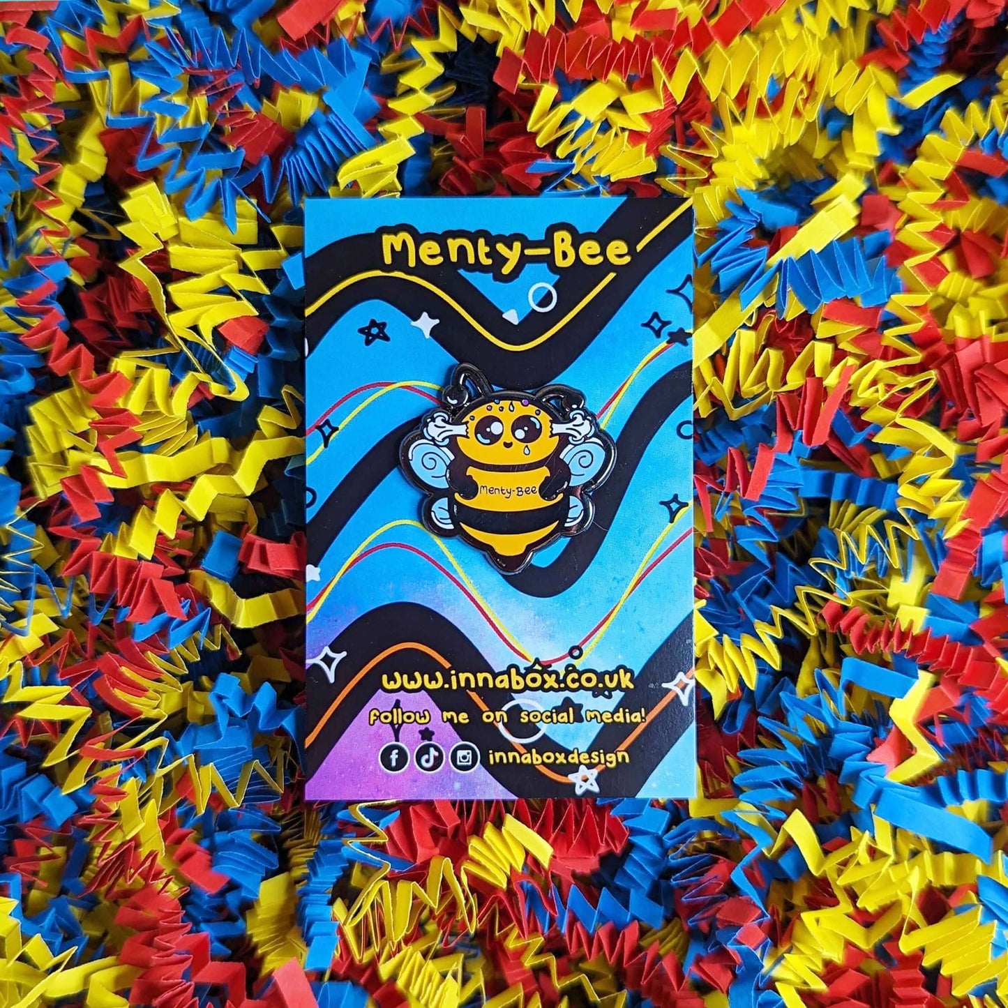 Menty-Bee Enamel Pin - Mental Breakdown pinned to its backing card with card confetti in the background. The enamel pin is of a bee looking stressed with steam coming out of its head and the text menty-Bee on its chest. Hand drawn design made to raise awareness for mental breakdowns