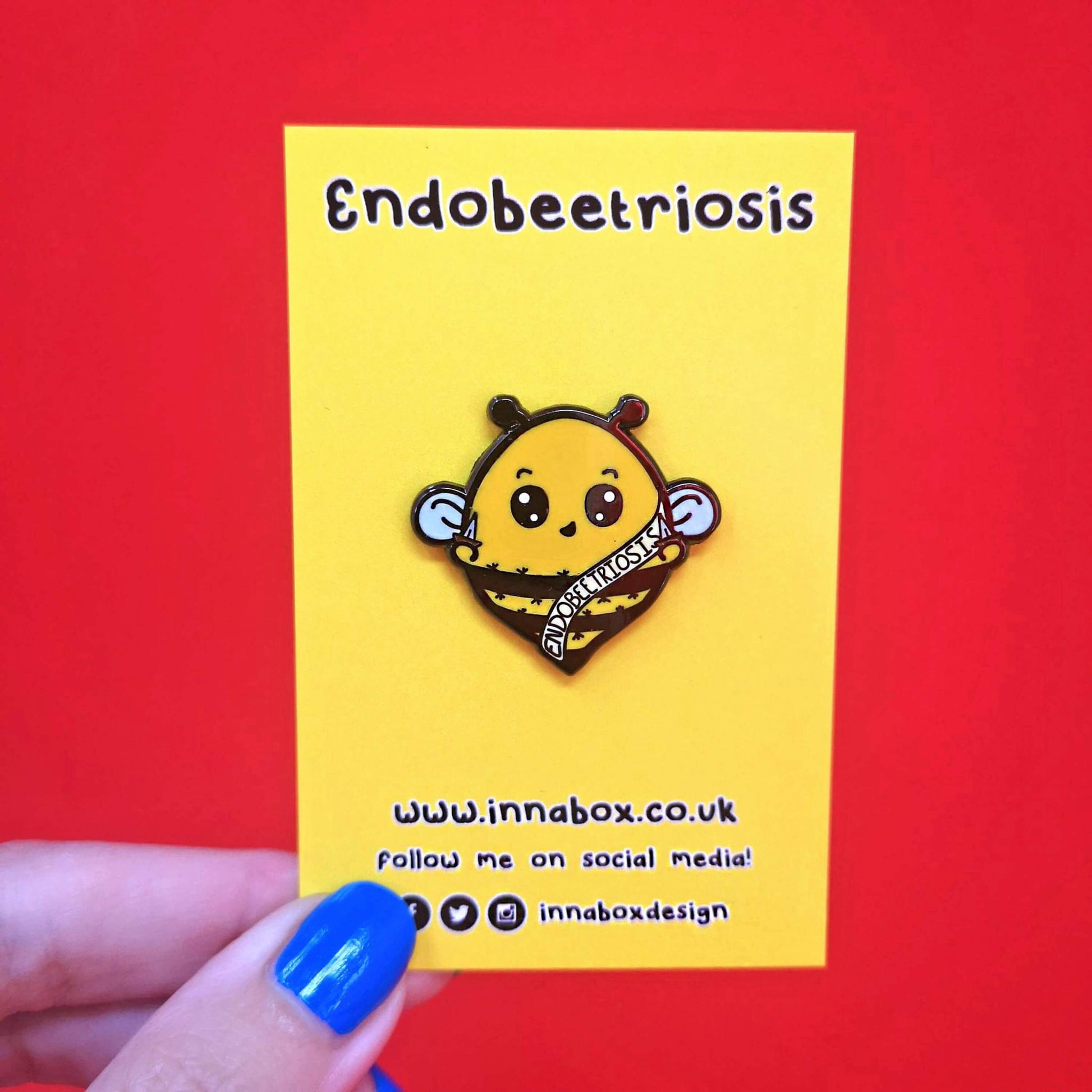 Endobeetriosis Enamel Pin - Endometriosis on yellow backing card on a red background. The enamel pin is a bumble bee with a smiley face and holding little daggers in its hands. The bee is wearing a white sash that has endobeetriosis written accross it. The enamel pin is designed to raise awareness for endometriosis