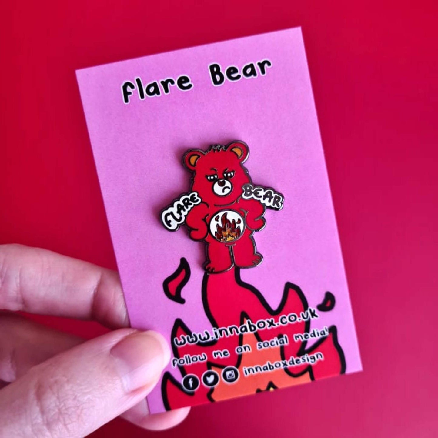 Flare Bear enamel pin on pink backing card with flames on held in front of a red background background. The enamel pin is of a red bear with a fed up expression and hands on its hips. There is a white circle on its belly with flames inside. Flare Bear is written on the pin. The enamel pin is designed to raise awareness for chronic illness flare ups.