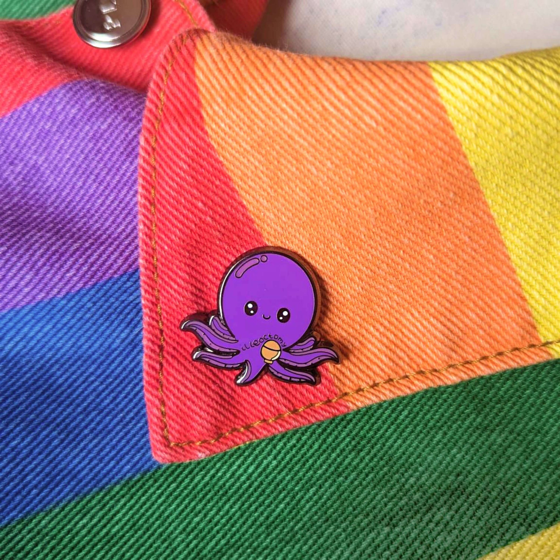 Ileoctomy Enamel Pin - Ileostomy shown pinned to a rainbow jacket. The enamel pin is a cute smiling purple octopus sticker with text saying ileoctomy on its belly with a stoma bag underneath. Enamel pin designed to raise awareness for Ileostomy