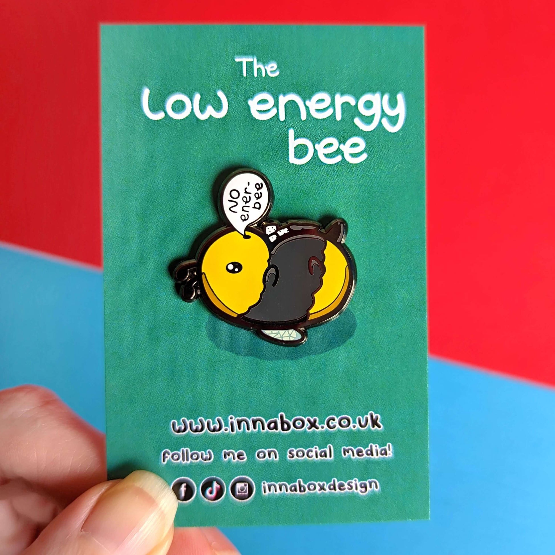 Low Energy Bee Enamel Pin on green backing held in front of a red and blue background. The enamel pin is a cute yellow and black bee lying on it's wings with its arms, legs, little belly and stinger in the air. The bee has a speech bubble coming from it's mouth with 'NO ener-bee' written inside in black writing. Hand drawn design made to raise awareness for chronic fatigue.