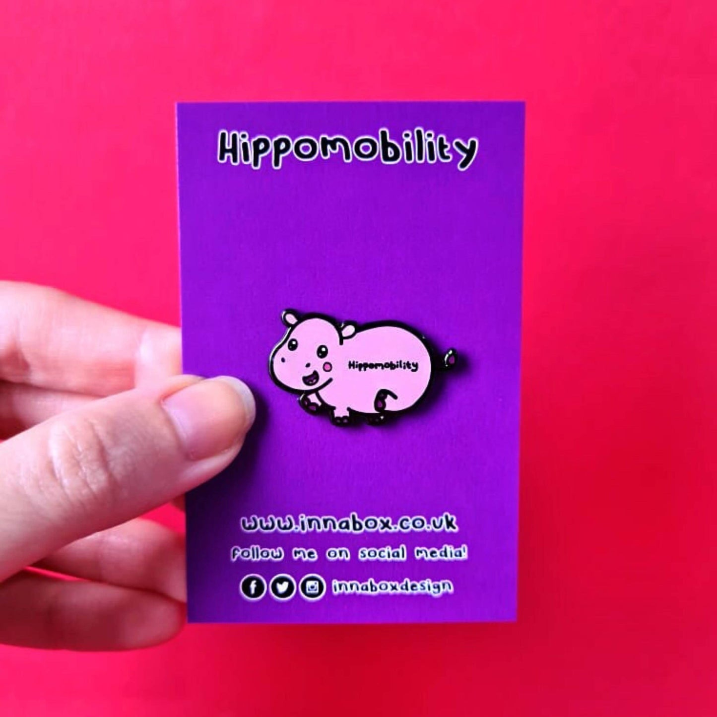 Hippmobility Enamel Pin - Hyper Mobility shown on purple backing card held in front of a red background. The enamel pin is of a pink happy hippo with the text hippomobility written on its stomach. The enamel pin is designed to raise awareness for hyper mobility