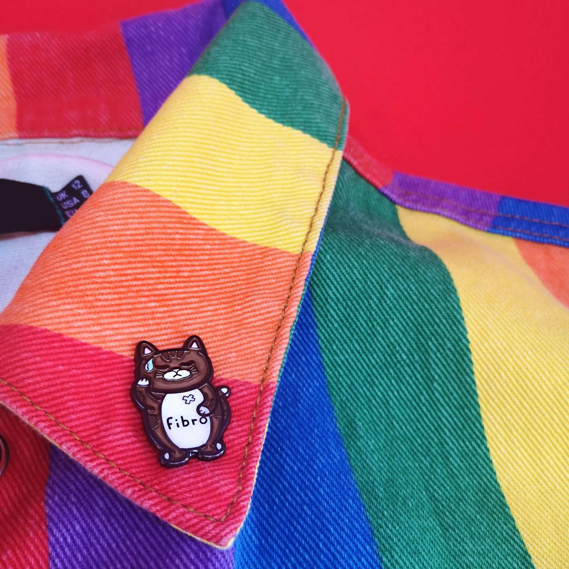 Fibromeowgia Enamel Pin - Fibromyalgia pinned to a rainbow coloured jacket. The enamel pin is a brown cat with a white stomach with fibro written across it. The cat has its eyes closed, a sweat droplet on its forehead and holding its head in its paw. The enamel pin is designed to raise awareness for fibromyalgia