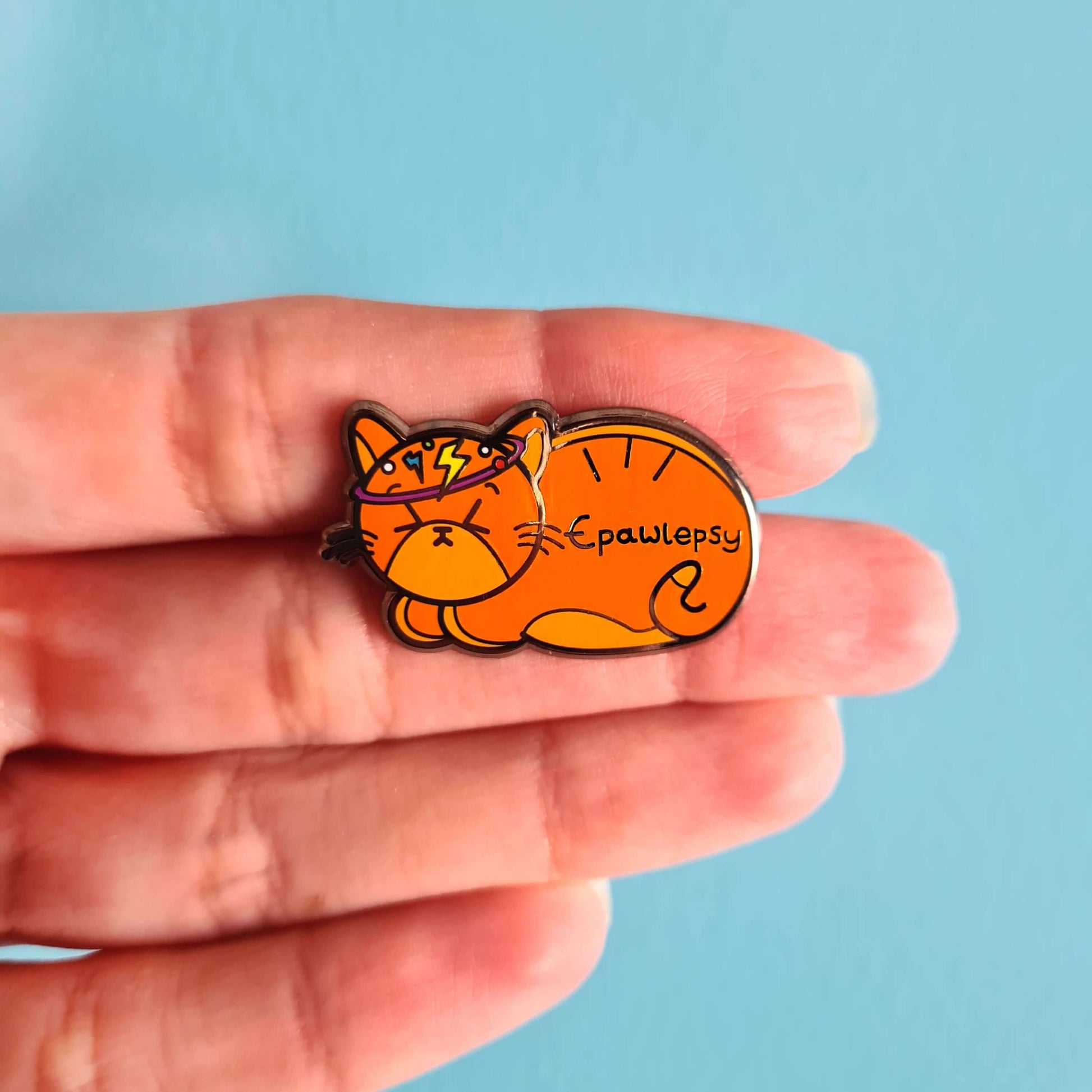Epawlepsy Enamel Pin - Epilepsy shown on palm of hand on a blue background. The enamel pin is a ginger cat with eyes scrunched closed and symbols to represent a dizzy spell drawn across his head. Epawlepsy is written on the cats stomach. Enamel pin is designed to raise awareness for epilepsy
