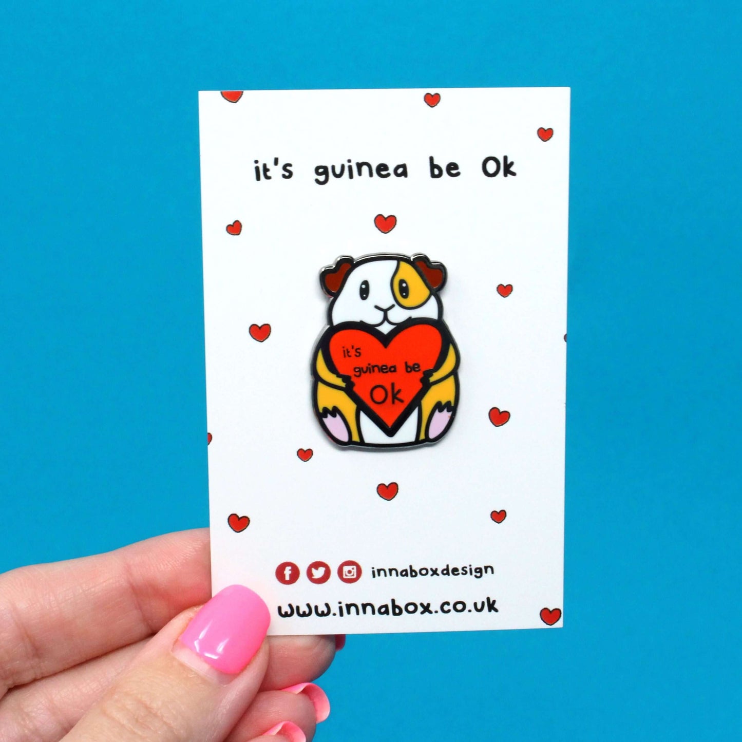 Guinea Pig Mental Health Enamel Pin on white backing card with red hearts held in front of a  blue background. The enamel pin is of a smiling guinea pig holding a big red heart with the text it's guinea be ok inside. The enamel pin is deigned to raise awareness for mental health.