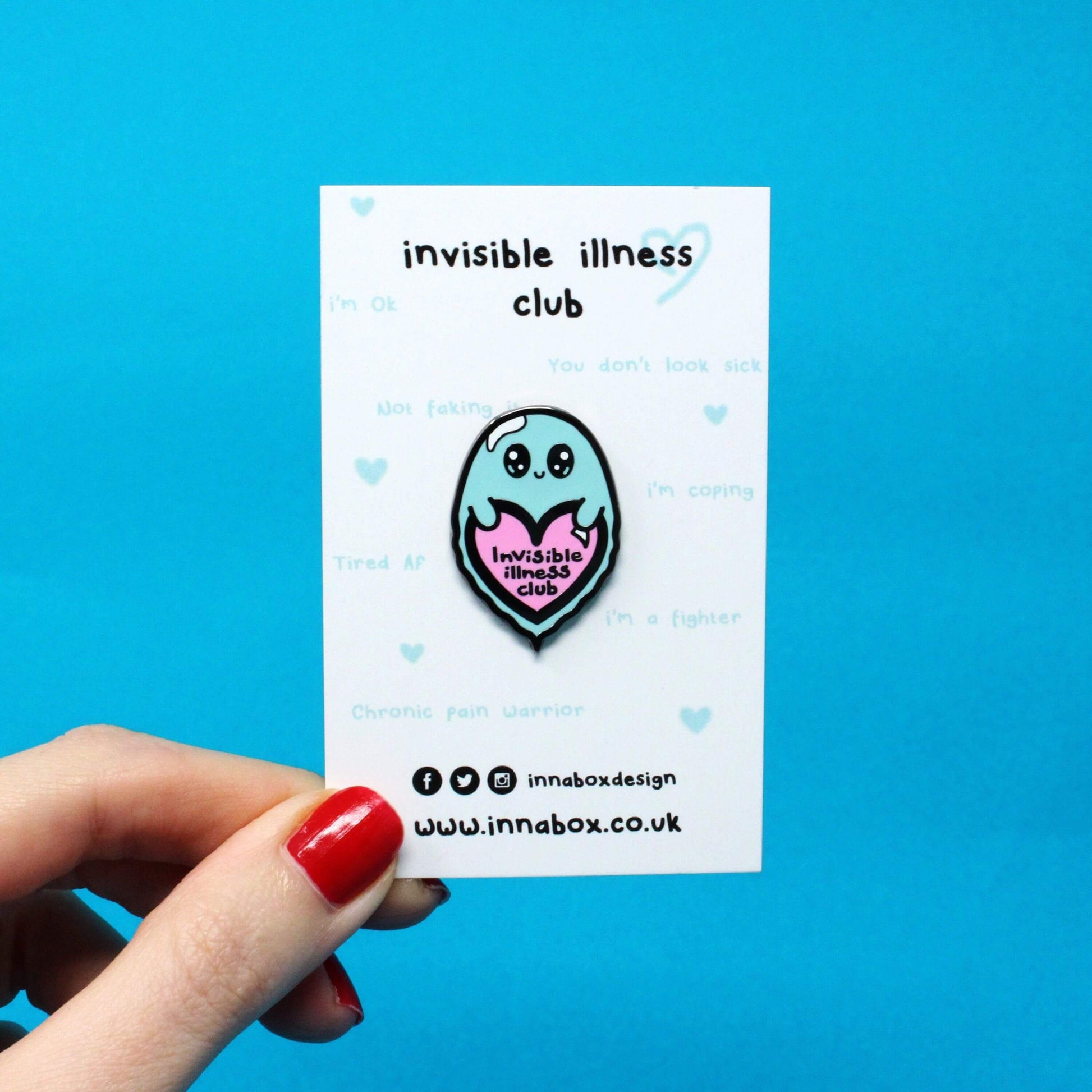 Invisible Illness Club Enamel Pin on white backing card with light blue quotes written on it in front of a red background. The enamel pin is of a cute smiling ghost holding a pink heart with text saying invisible illness club. The enamel pin is designed to raise awareness for hidden and chronic illnesses