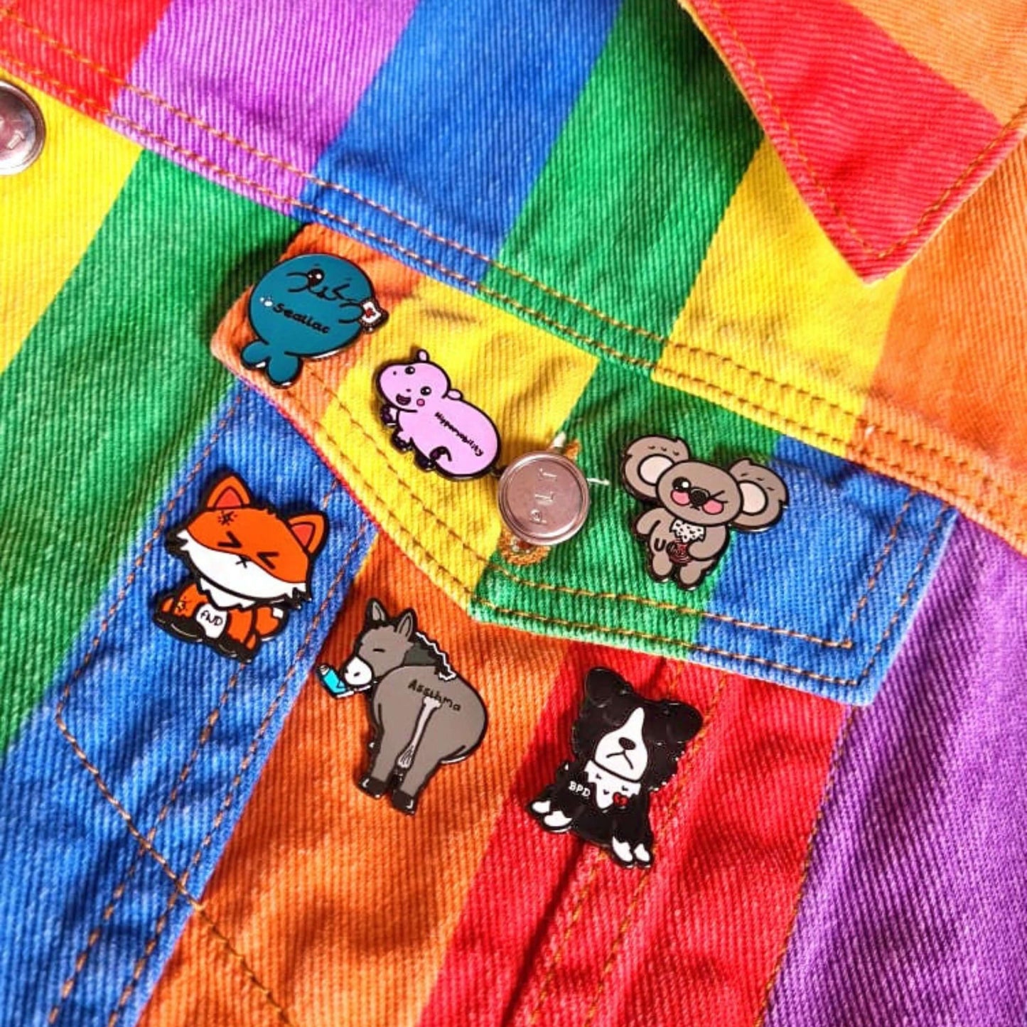multiple Innabox enamel pins pinned onto a rainbow coloured jacket. The enamel pins are designed to raise awareness for chronic illnesses 